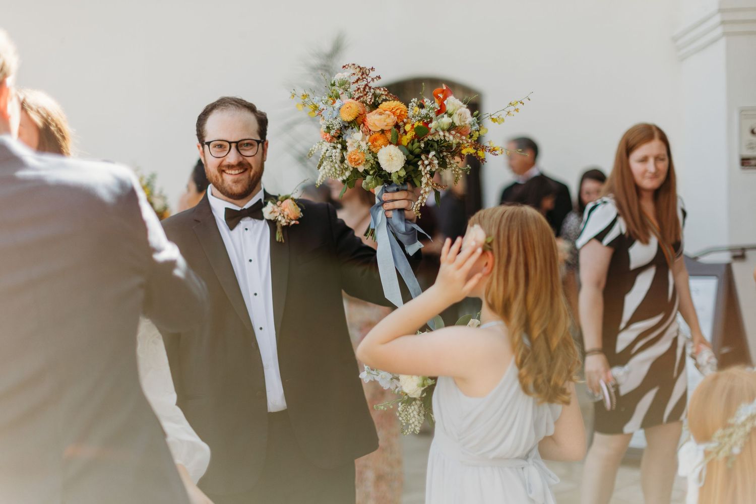 Wedding celebration candid moment with floral bouquet and guests in formal attire outside SLO Mission.