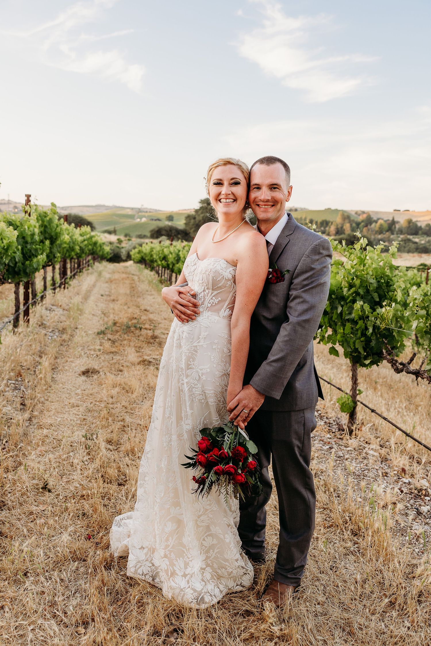 A happy couple poses together in a vineyard setting holding a red floral bouquet.