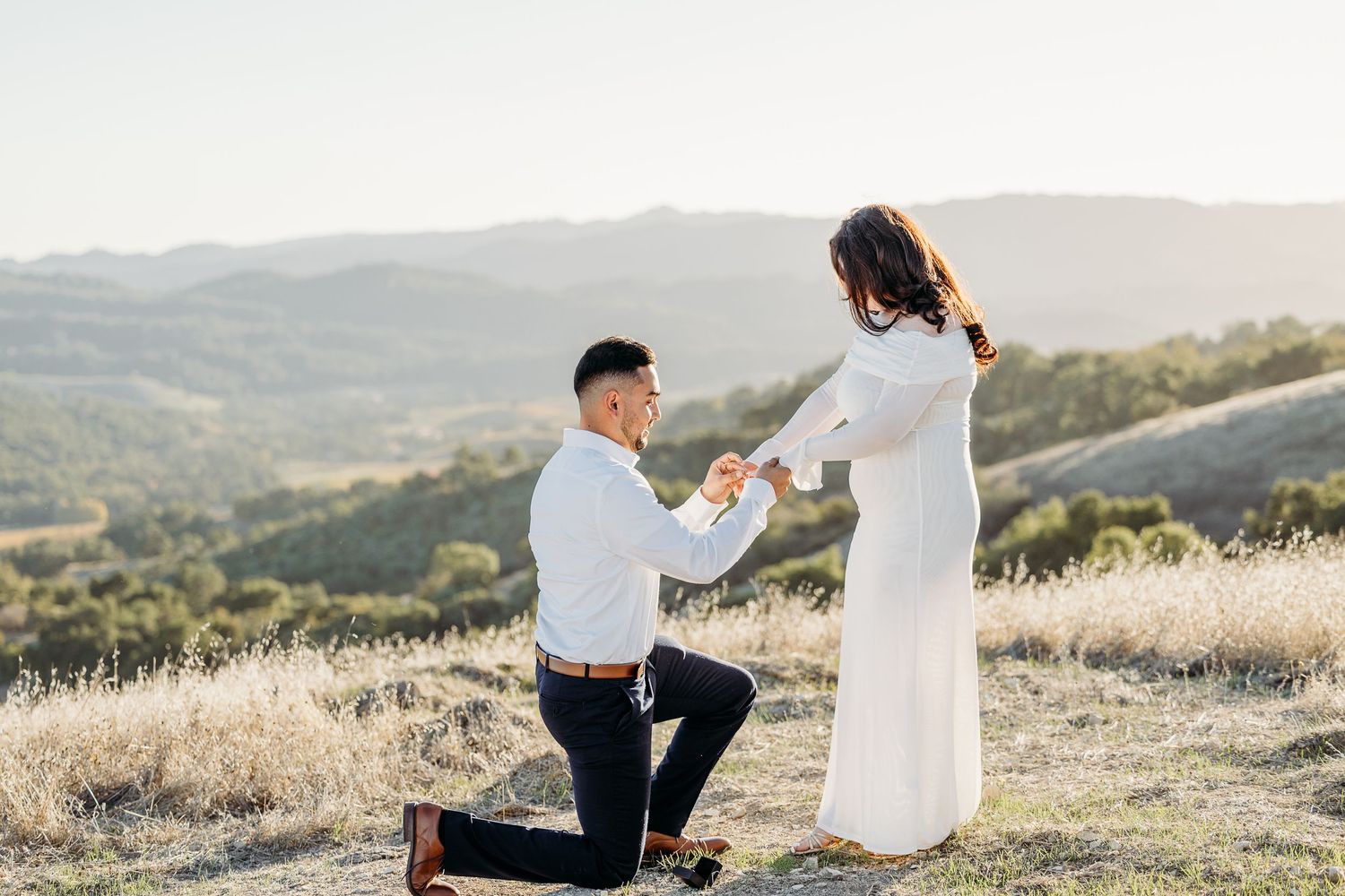 A couple shares a romantic moment during sunset on a hilltop overlooking rolling countryside.