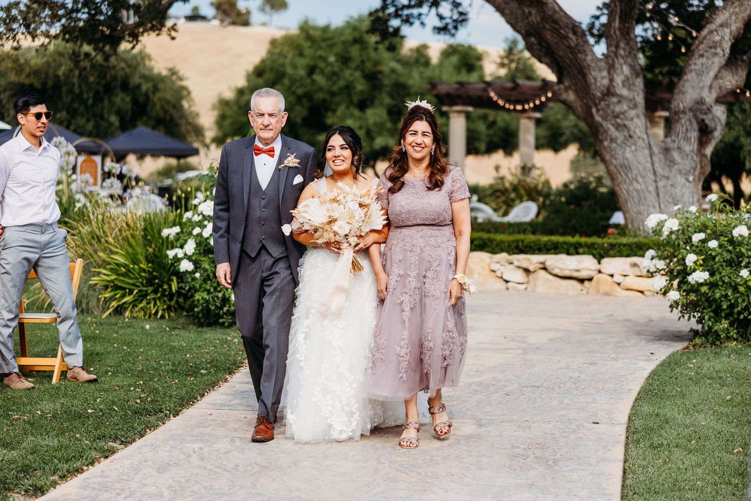 Wedding party members walk down stone path during outdoor garden ceremony.
