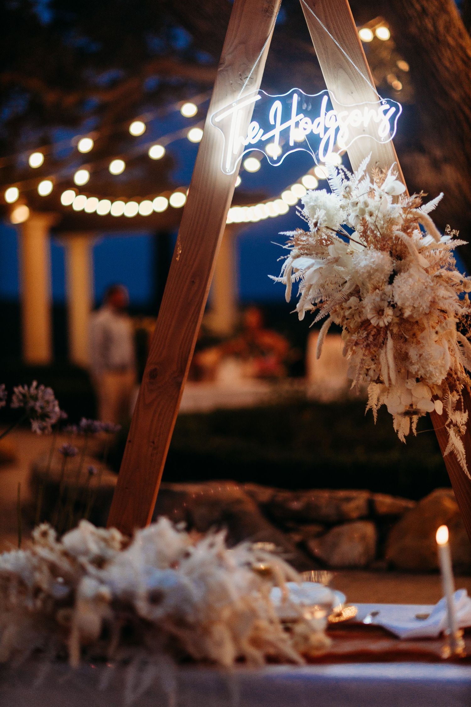 Elegant wedding decor featuring dried floral arrangements and neon signage under string lights at dusk.