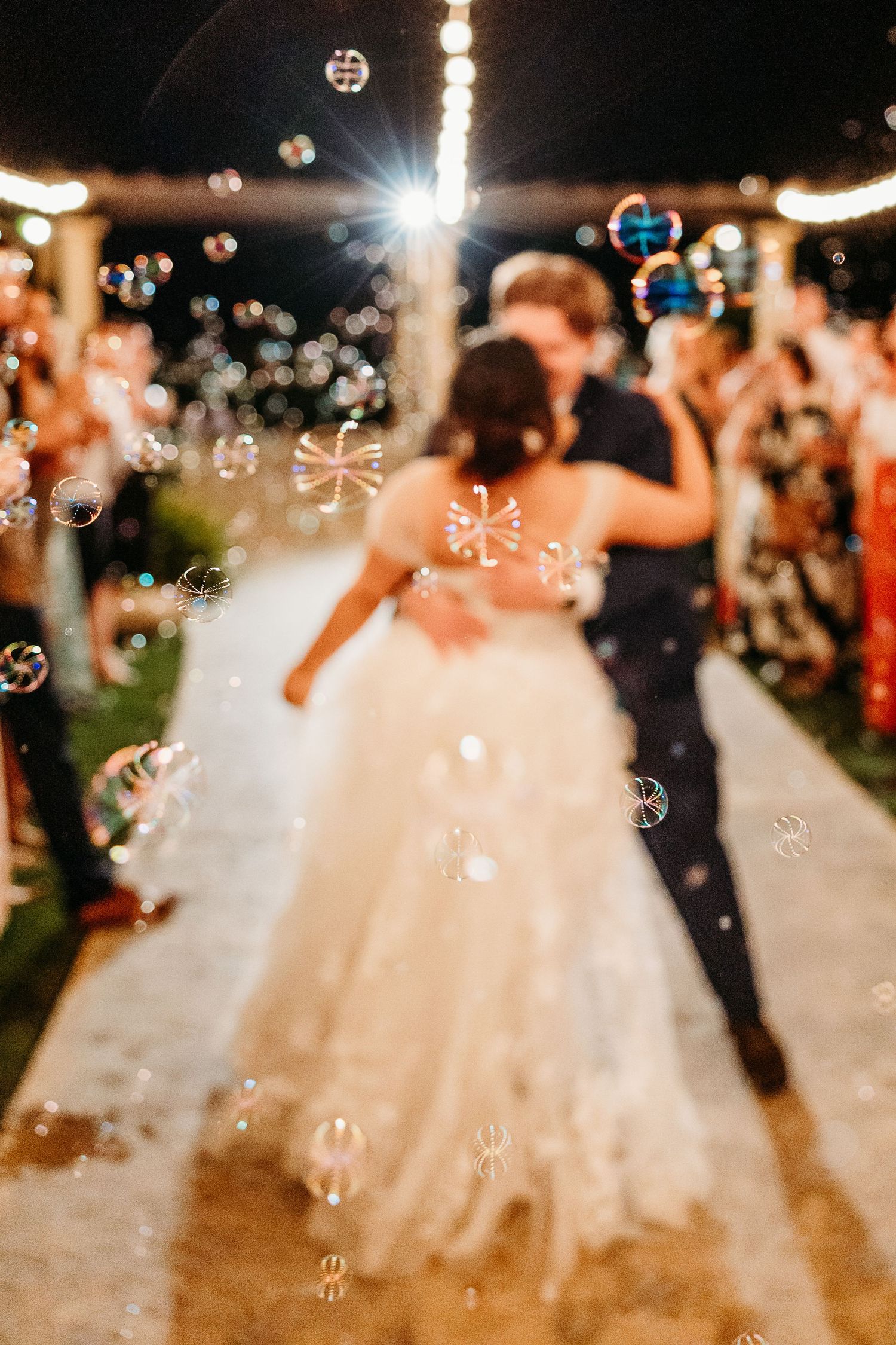 Dancing couple surrounded by floating bubbles under string lights at outdoor reception.