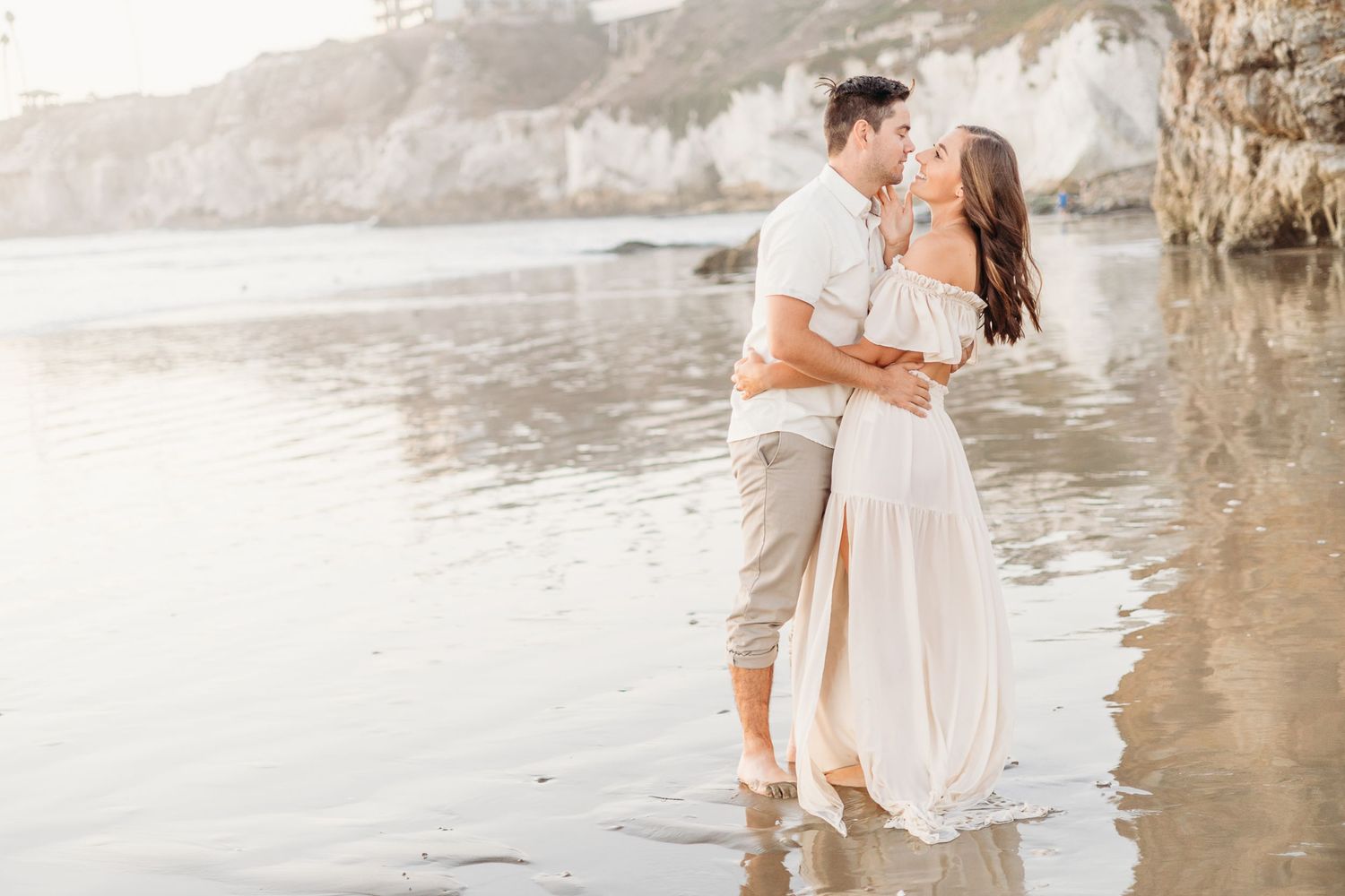 Couple in light summer clothes sharing tender moment on beach with cliffs in background.