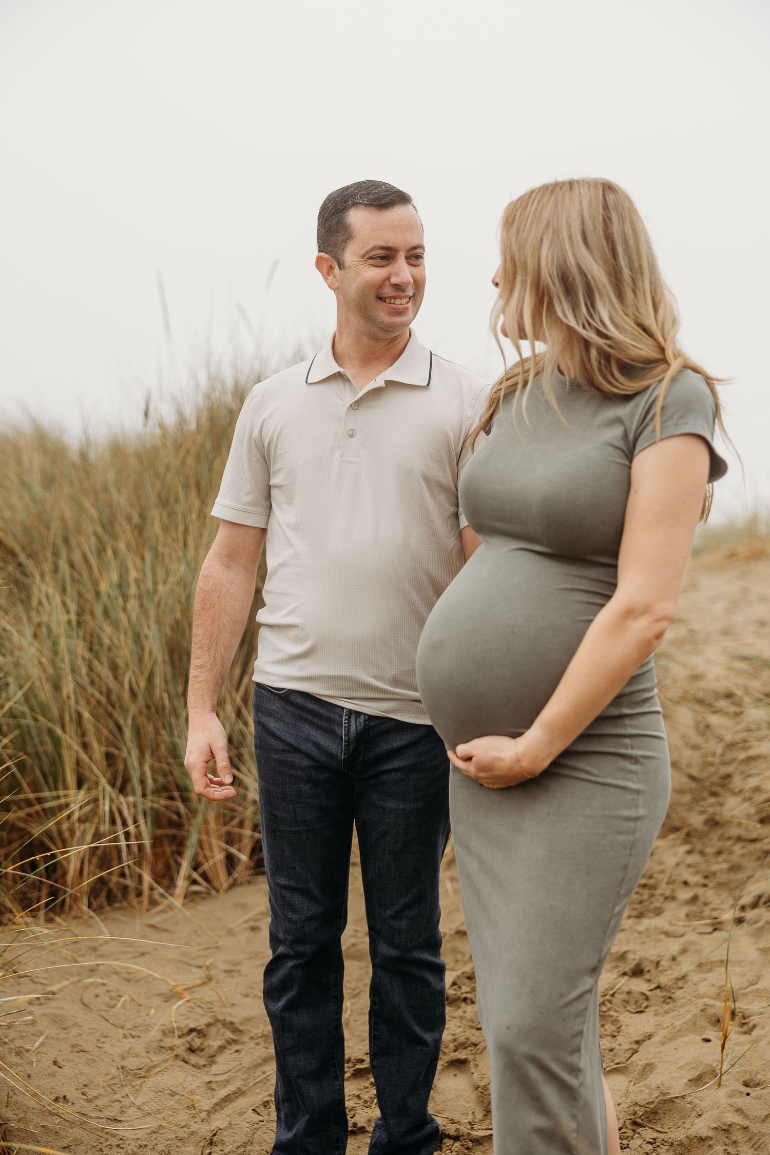 An expecting couple shares a loving moment while standing on sandy coastal terrain.