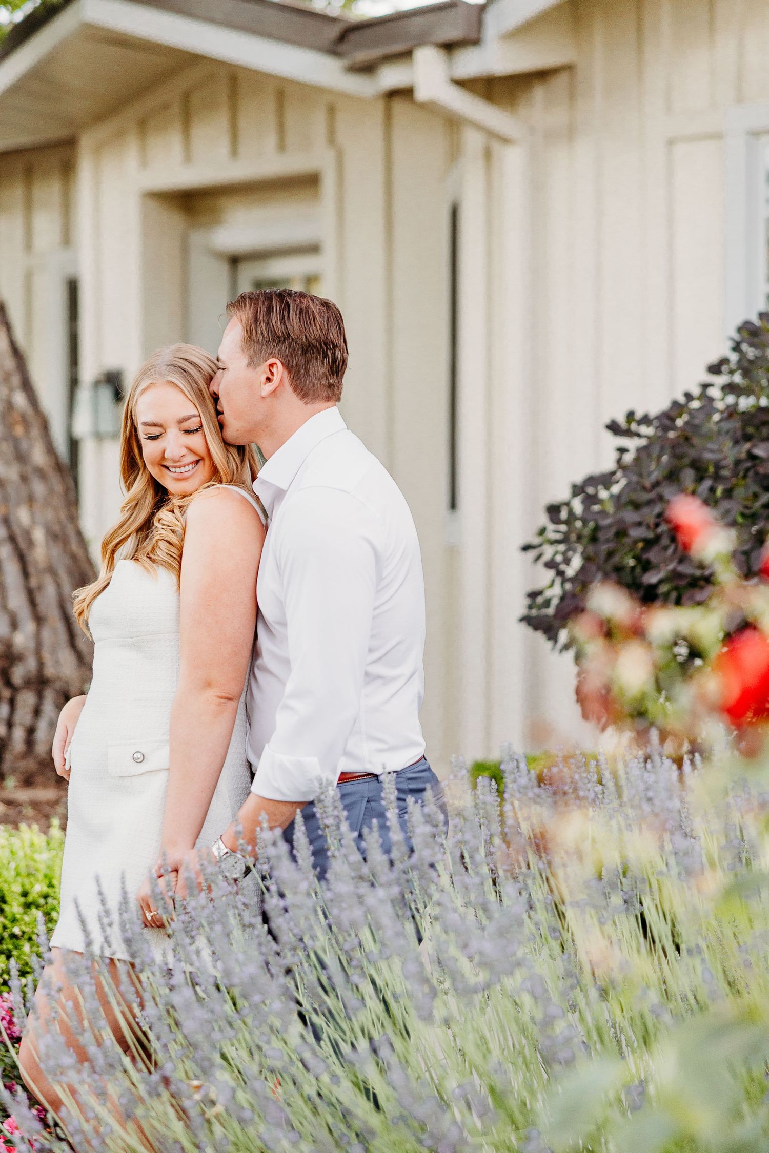 A couple embraces in a garden with blooming lavender flowers and white building in the background.