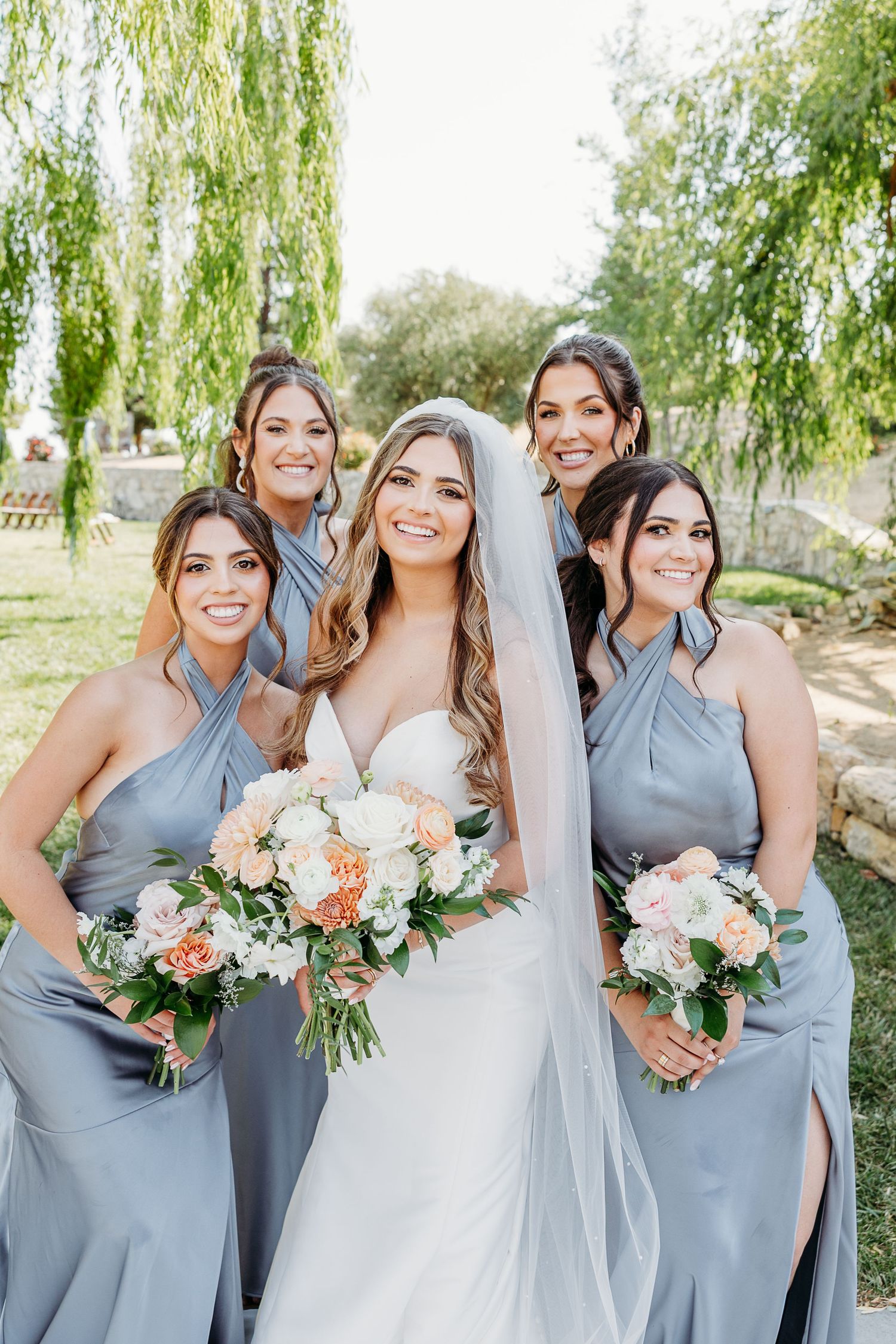 Wedding party wearing light blue bridesmaid dresses and white bridal gown posing under willow trees with floral bouquets.
