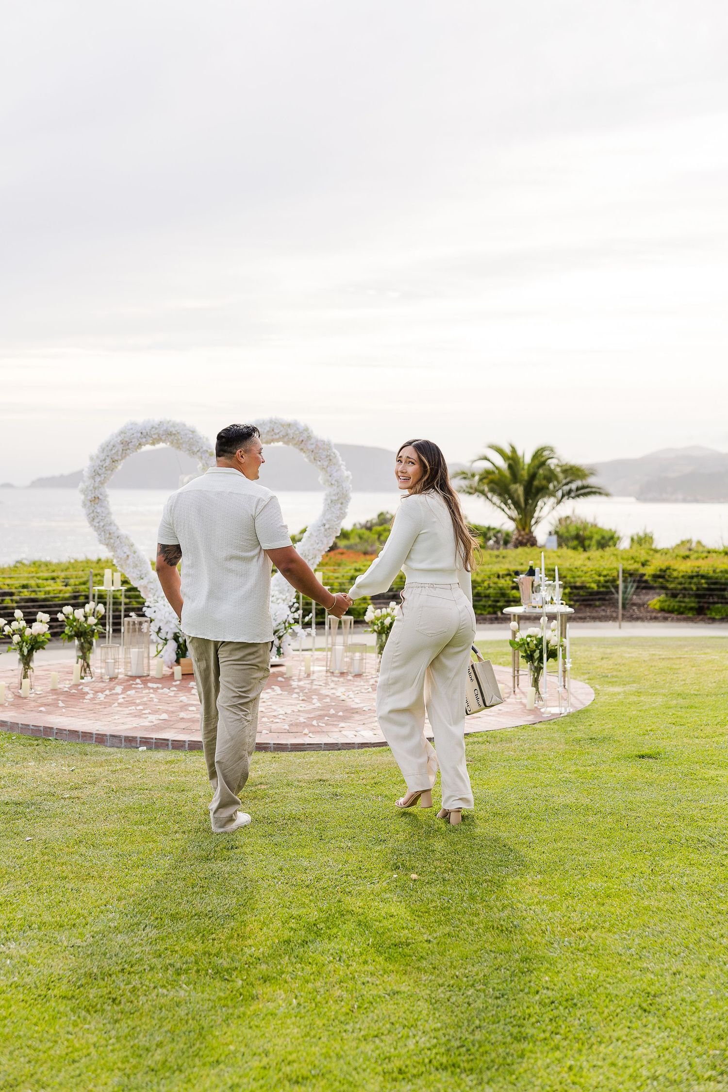 A couple in white outfits dancing playfully on a grassy lawn with palm trees and mountains in the background.