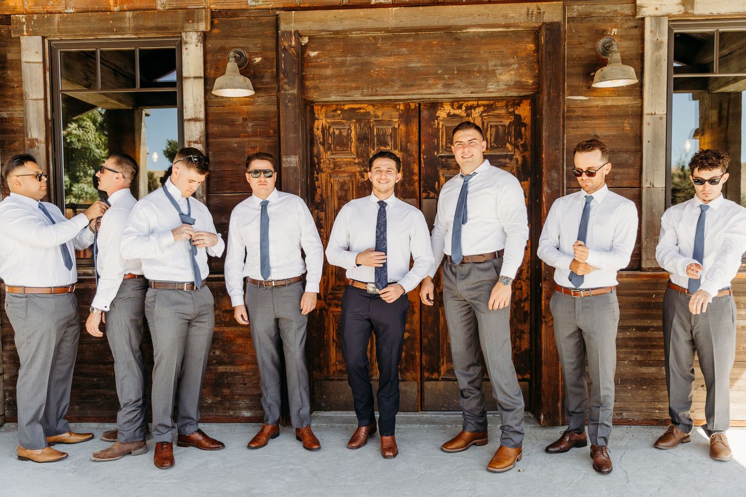 Groomsmen lined up in gray suits and blue ties pose casually against a rustic wooden backdrop.