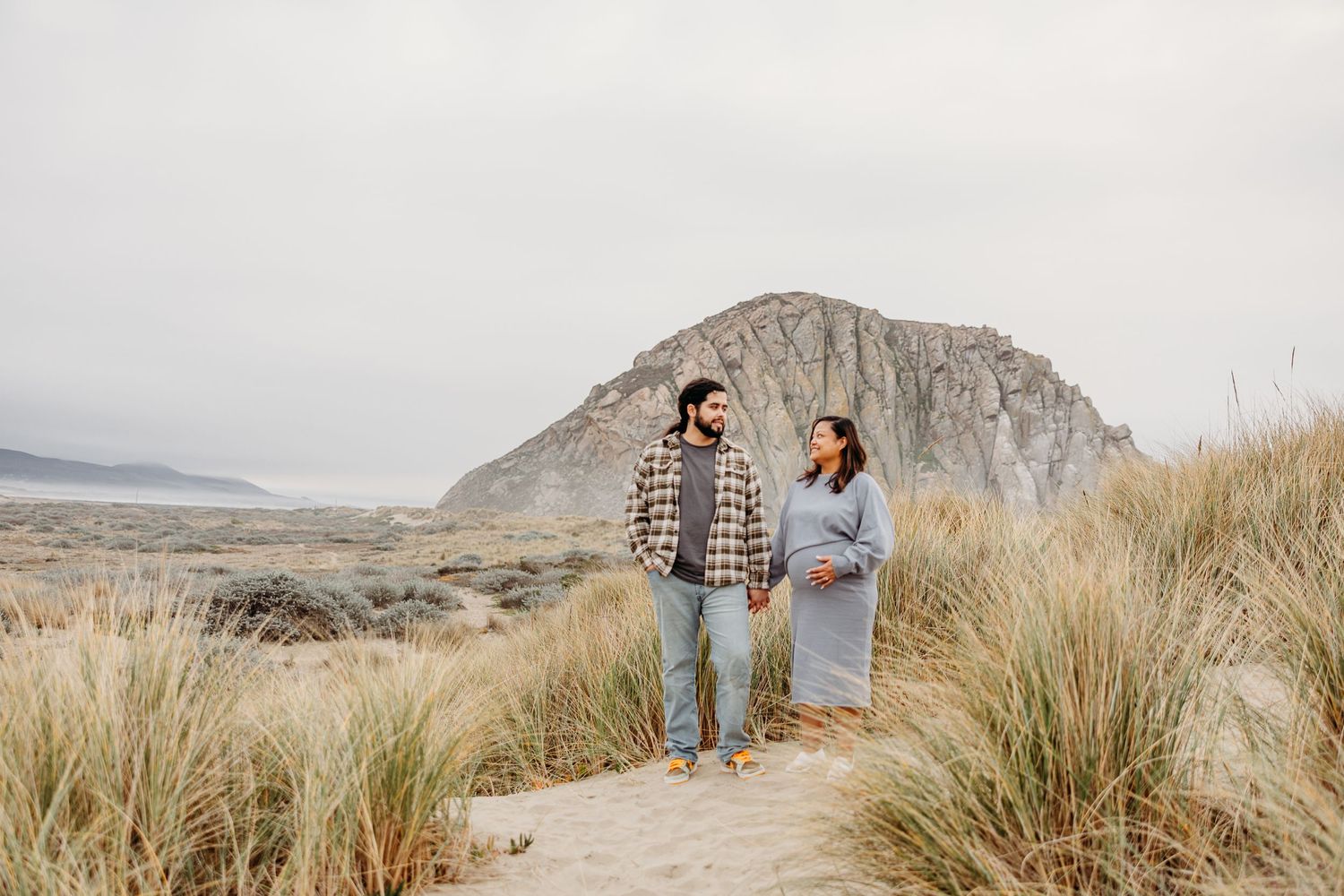 Couple stands together on mountain trail with scenic desert landscape and large rock formation in background.