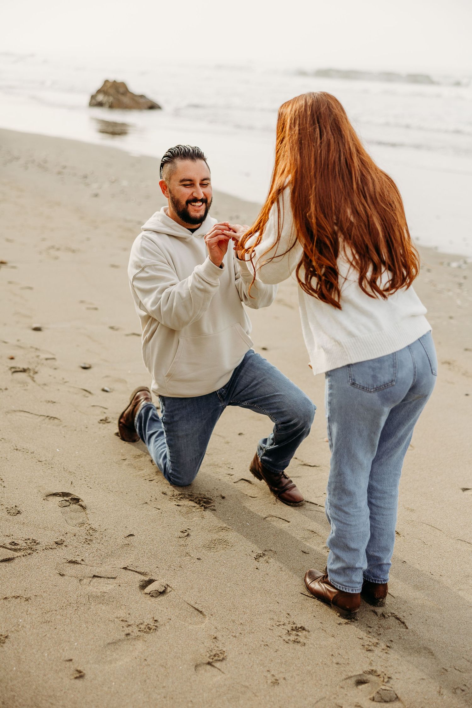 A person proposes on one knee to another on a misty beach with waves crashing in the background.
