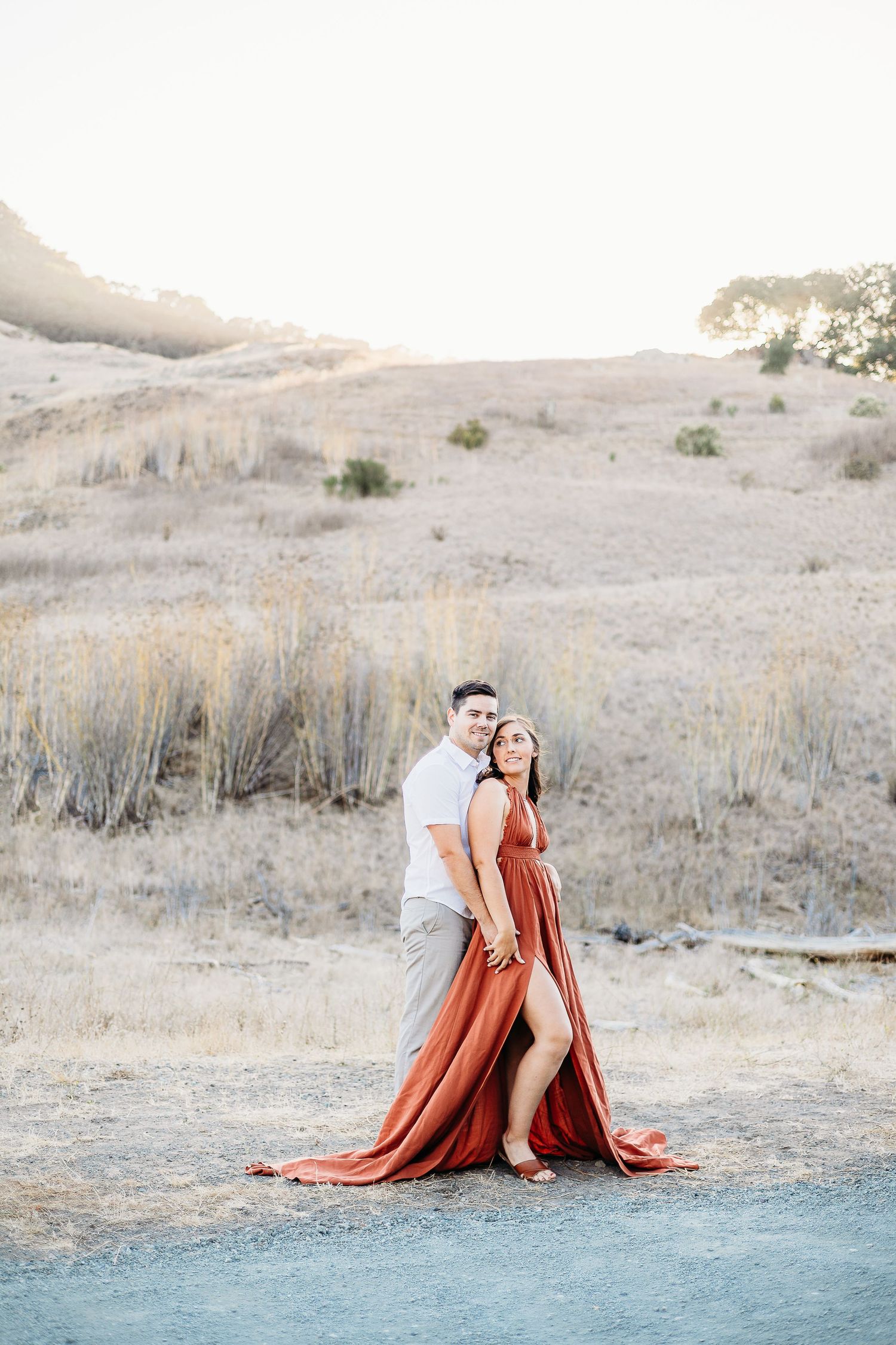 A couple stands together on a dirt path with rolling hills behind them at sunset.