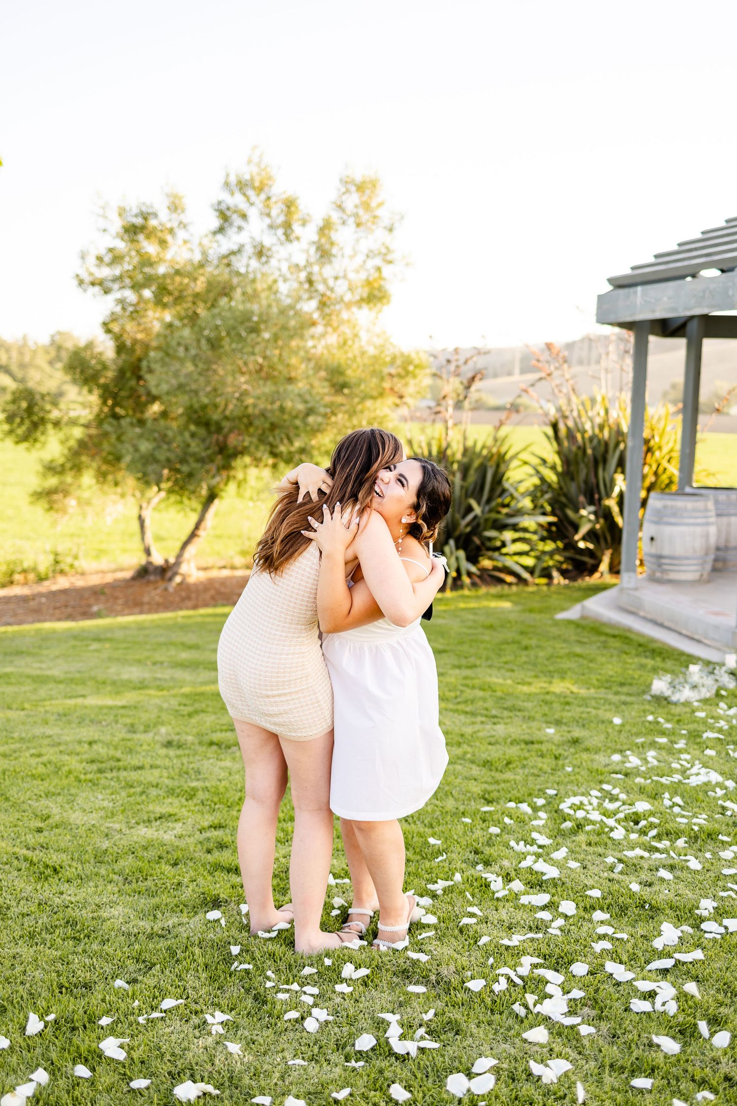 Two people share an emotional embrace on grass covered in white flower petals during golden hour.