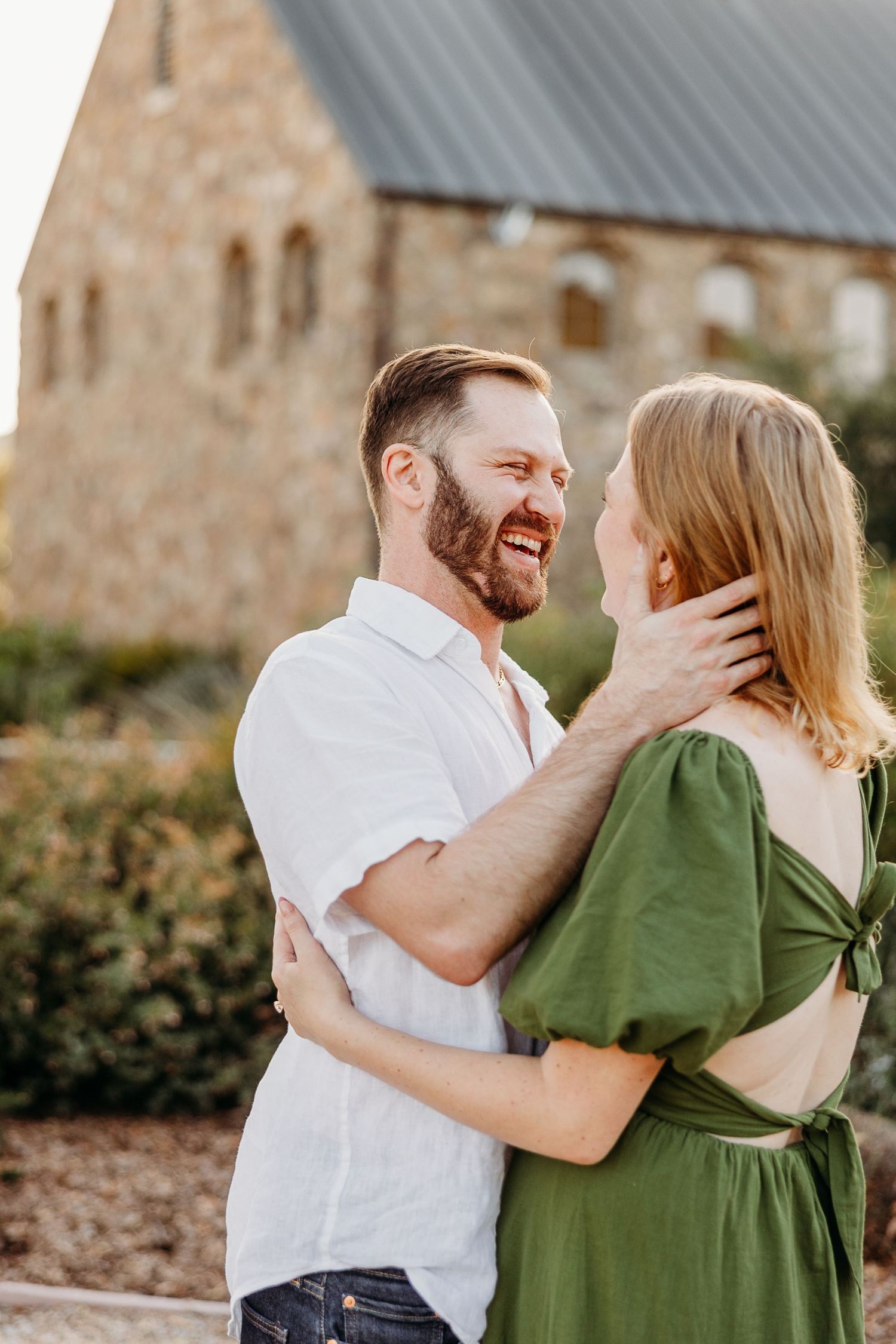 A romantic couple in white and green outfits embrace and laugh together during an outdoor photo session.