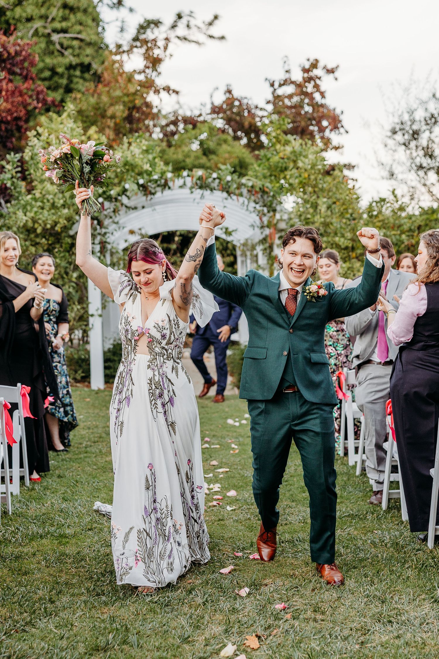 Wedding party celebrates joyfully on a grassy lawn with guests throwing confetti and cheering.