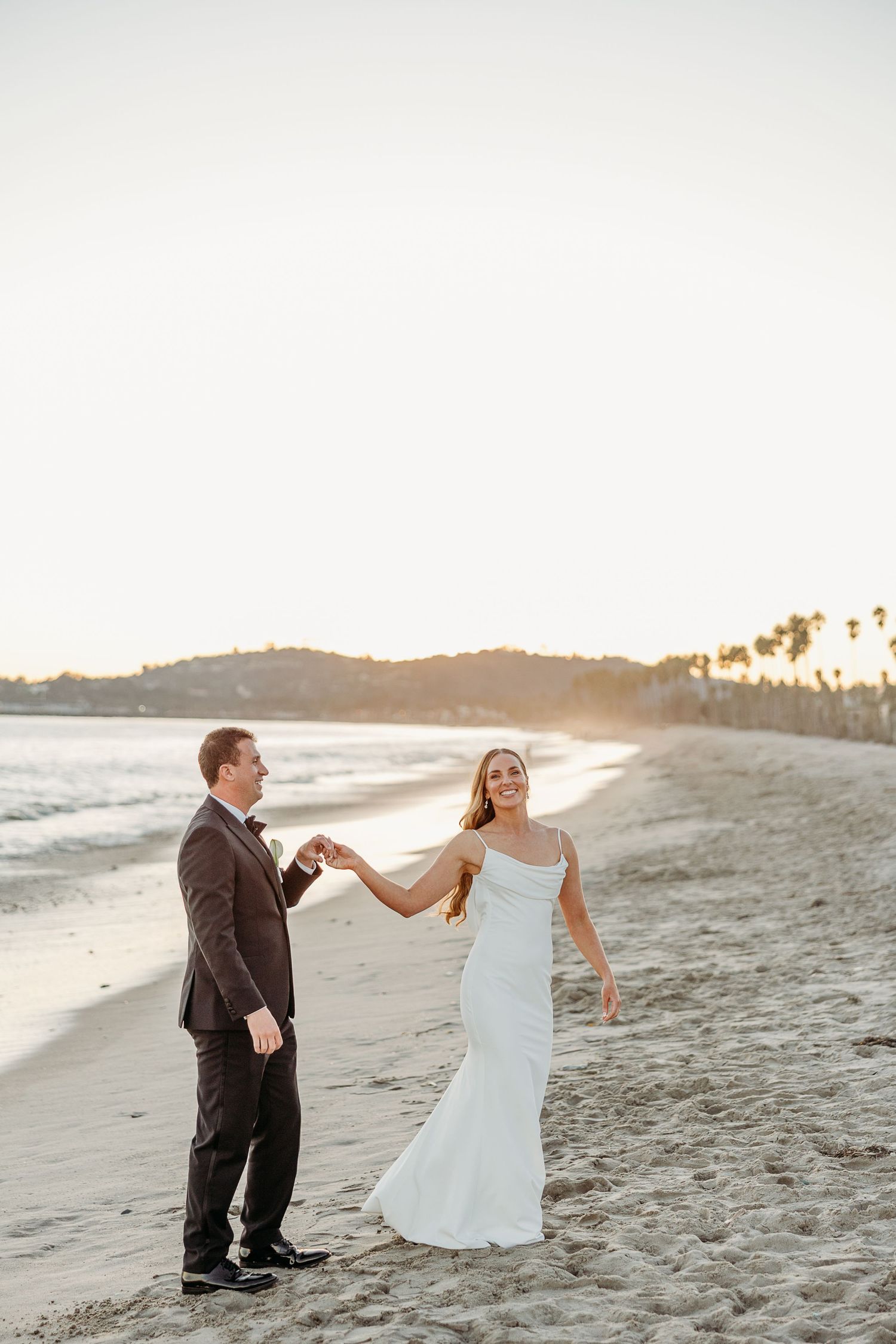 A couple dances on the beach at sunset with palm trees and ocean waves in the background.