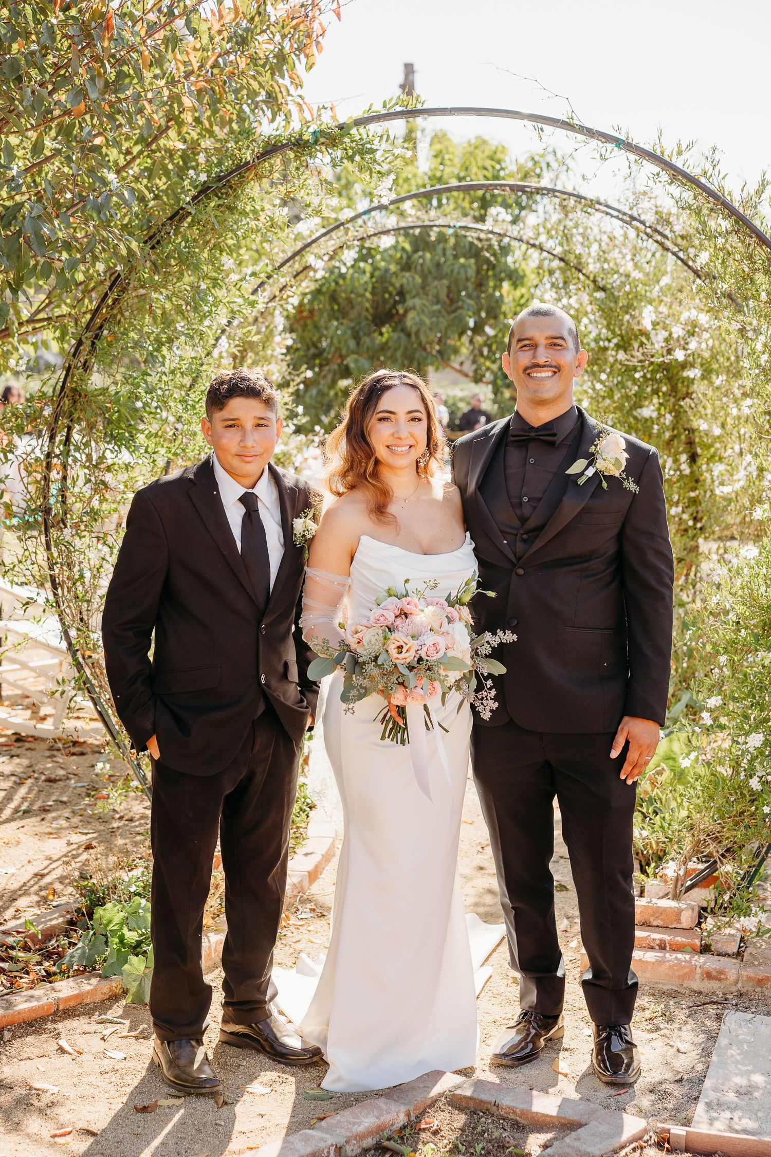 A wedding party poses under a floral arch in a garden setting wearing formal black suits and a white bridal gown.