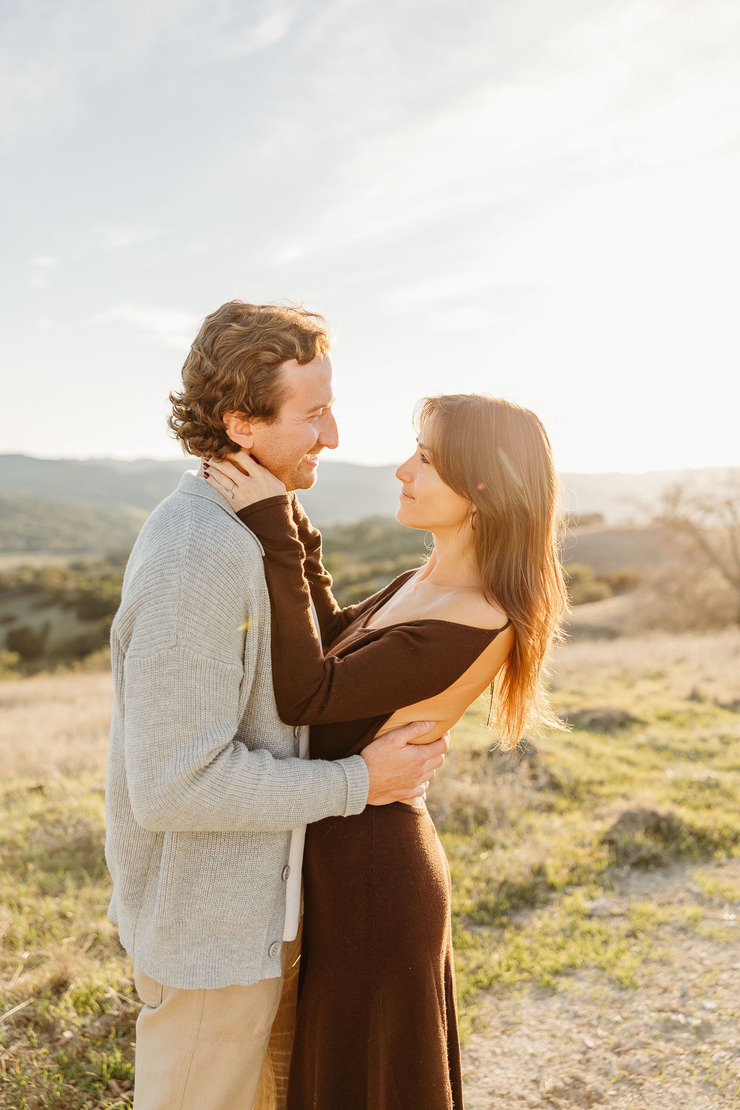 A couple embraces on a hilltop during golden hour with scenic mountain views in the background.