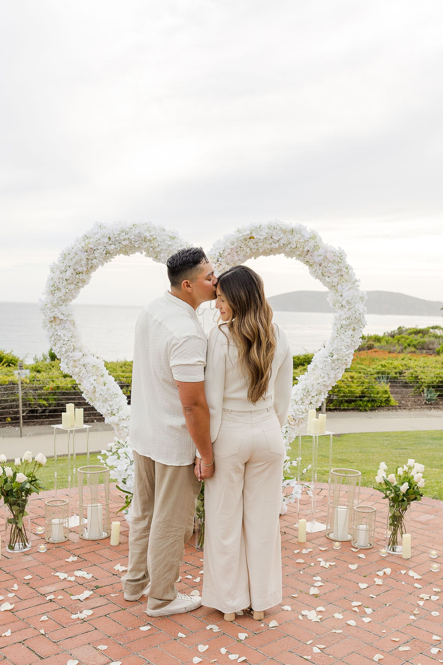 A couple shares a romantic moment under a white floral heart-shaped arch overlooking the ocean at an outdoor ceremony.