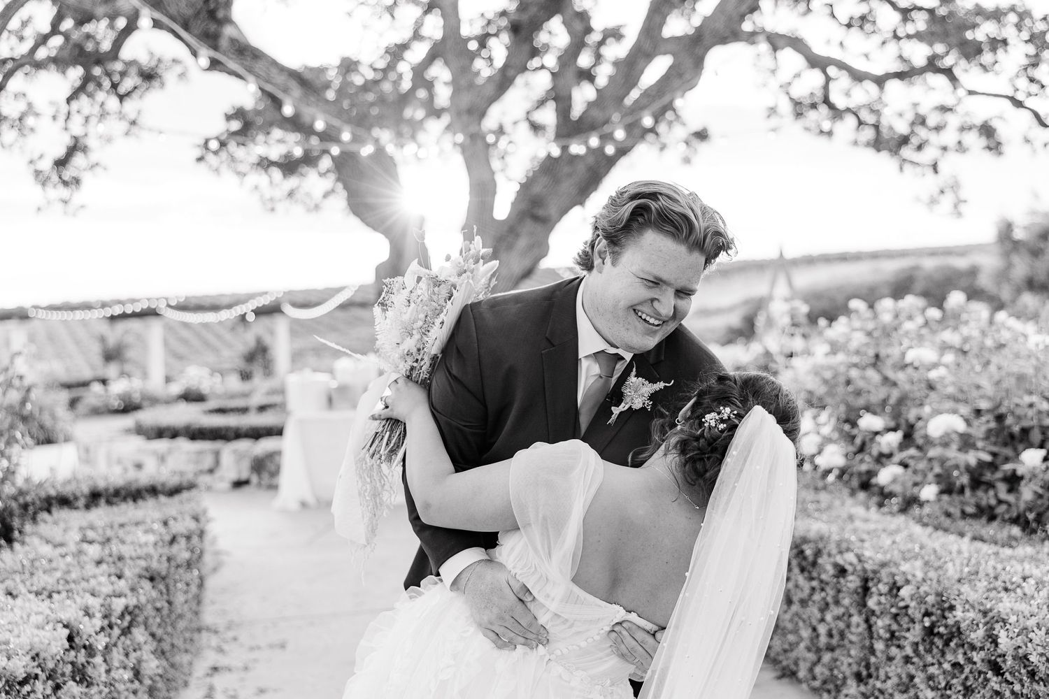 A black and white wedding photo captures a romantic moment in a garden setting.