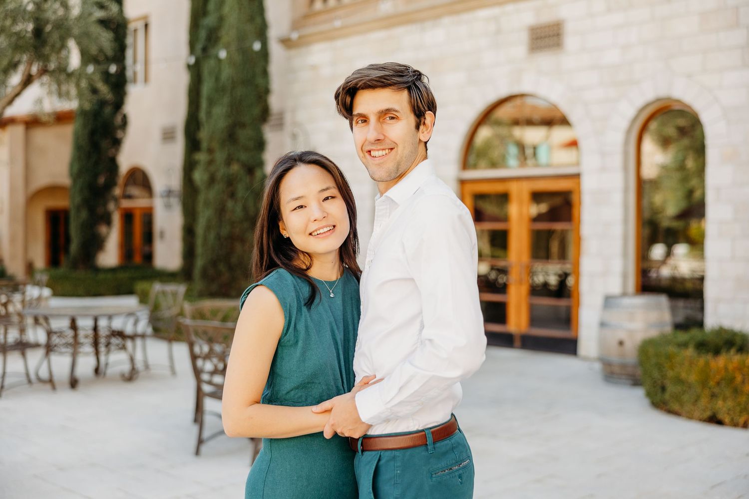 A couple embraces and smiles while standing in front of an elegant arched building entrance.