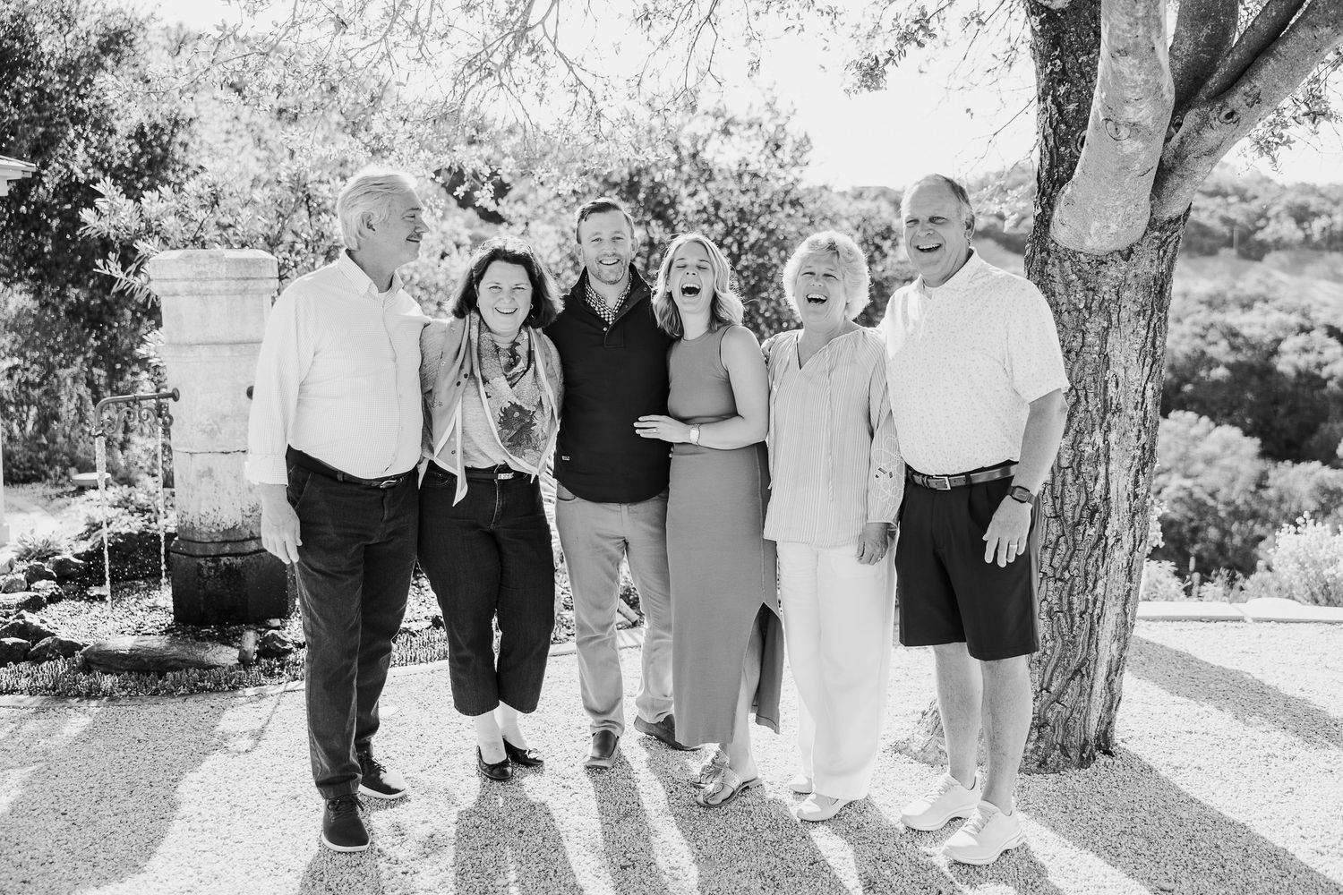 A group of people stand together outdoors in a casual gathering under trees on a sunny day.