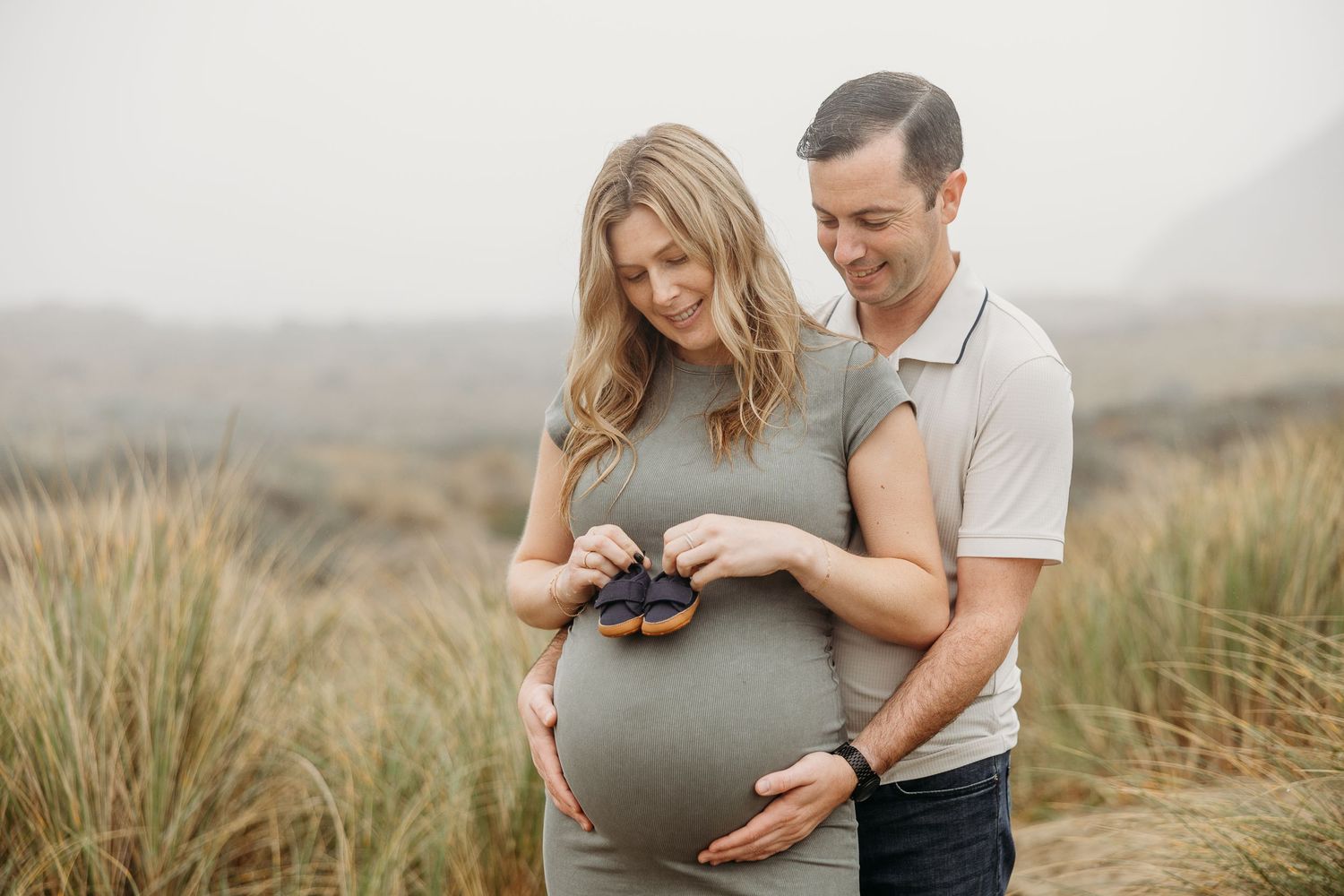 Couple shares intimate moment outdoors while holding baby shoes against pregnant belly.