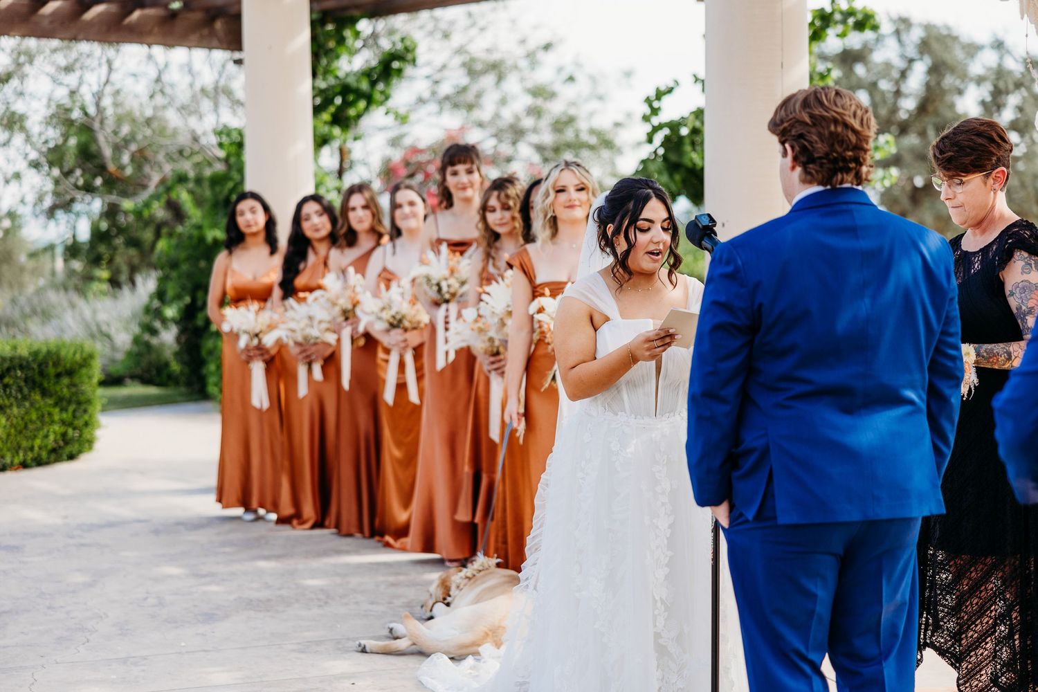 Bridesmaids in rust-colored dresses stand in line during outdoor wedding ceremony.