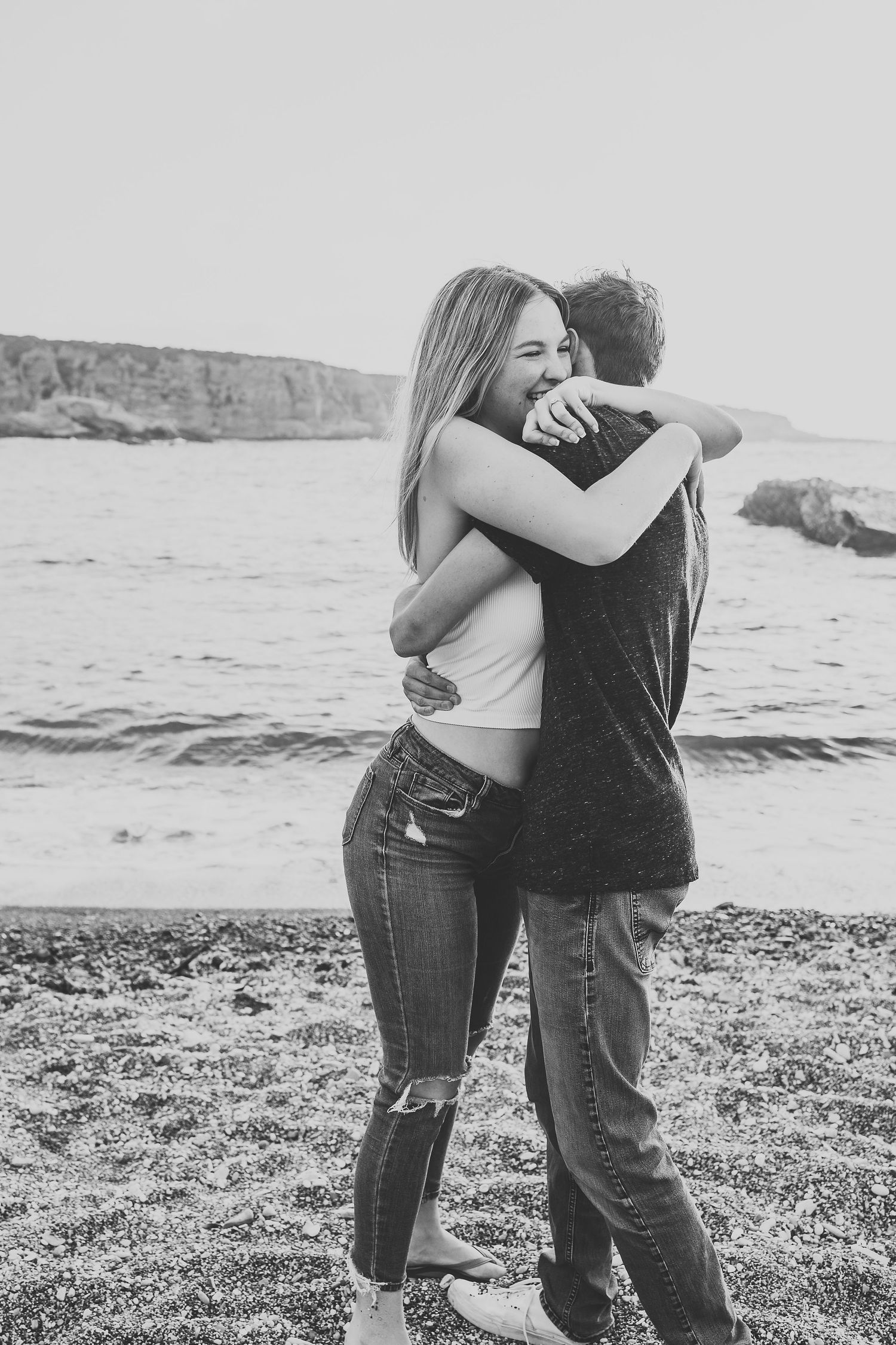 A couple embraces on a beach with rocky cliffs in the background during a black and white photoshoot.