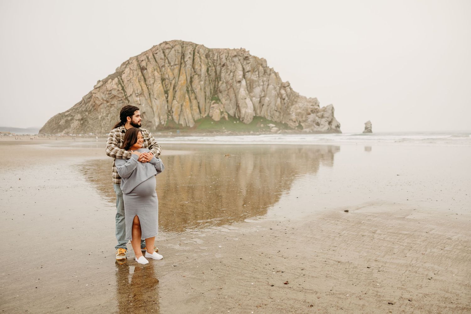 Someone stands on a misty beach with iconic Morro Rock formation visible in the background at California's central coast.