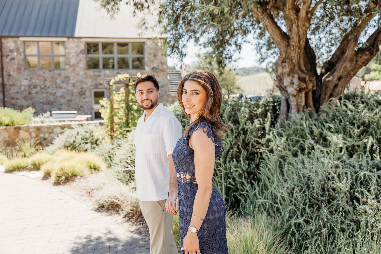 A couple poses together in a garden setting with green foliage and stone architecture in the background.