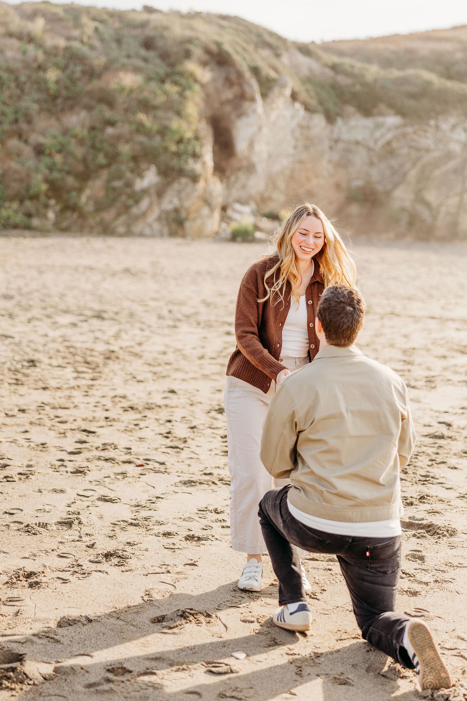 A couple shares a romantic moment on a sandy beach with cliffs in the background during golden hour.