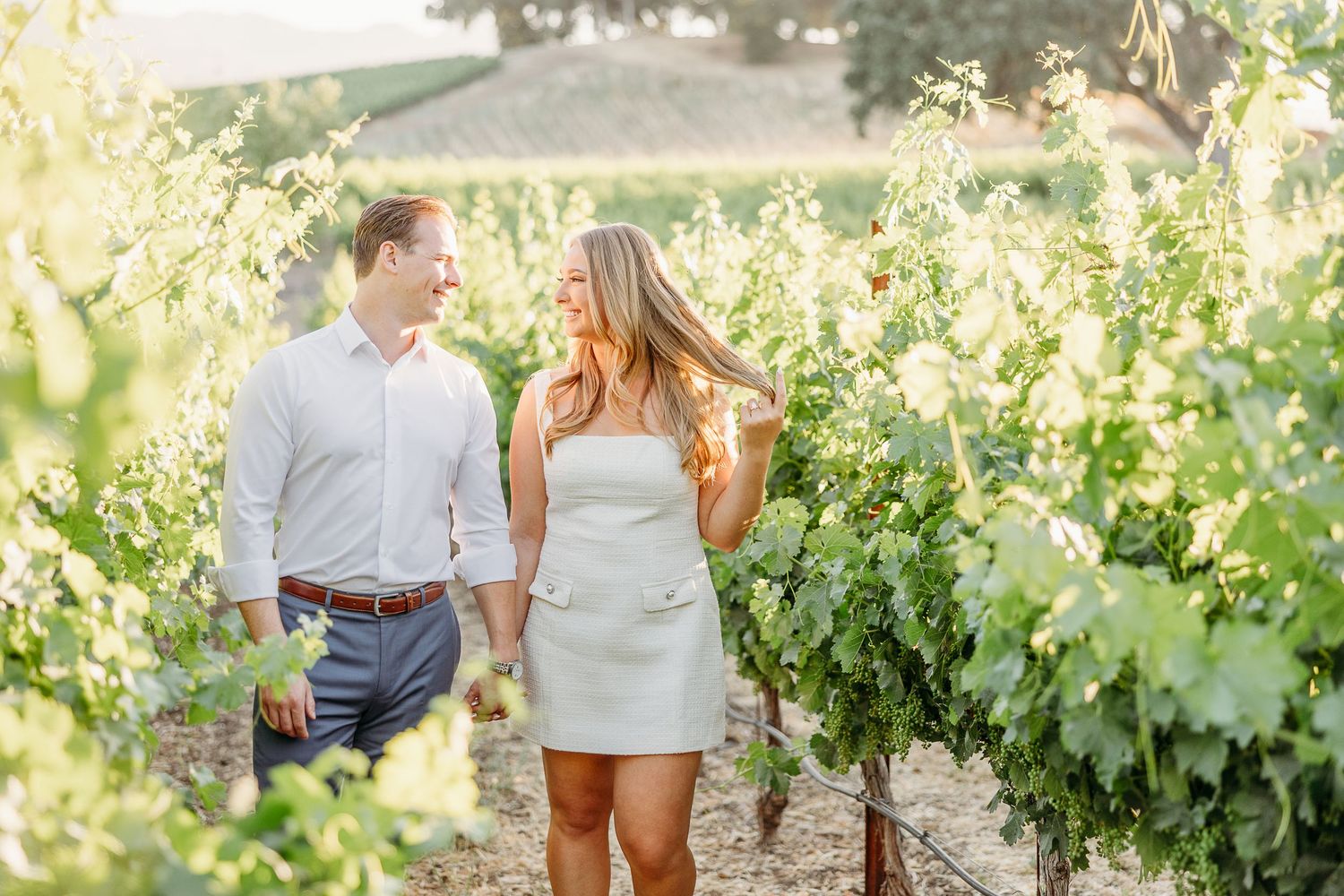A couple walking through lush green vineyard rows during sunset in wine country.
