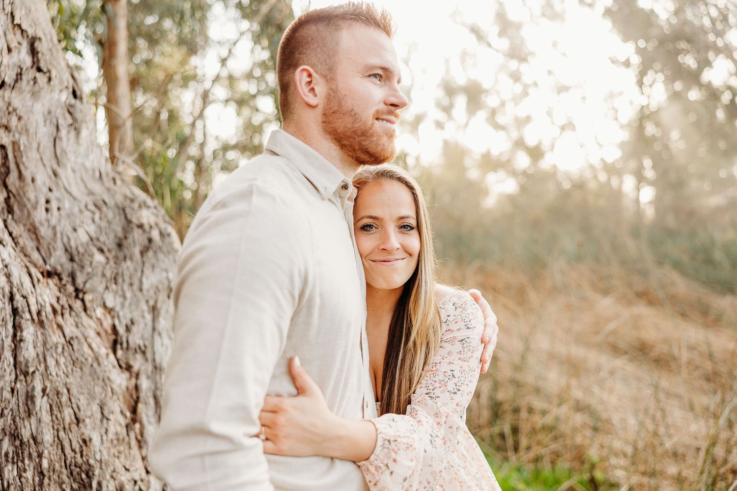 A couple embraces against a tree trunk in soft natural lighting.