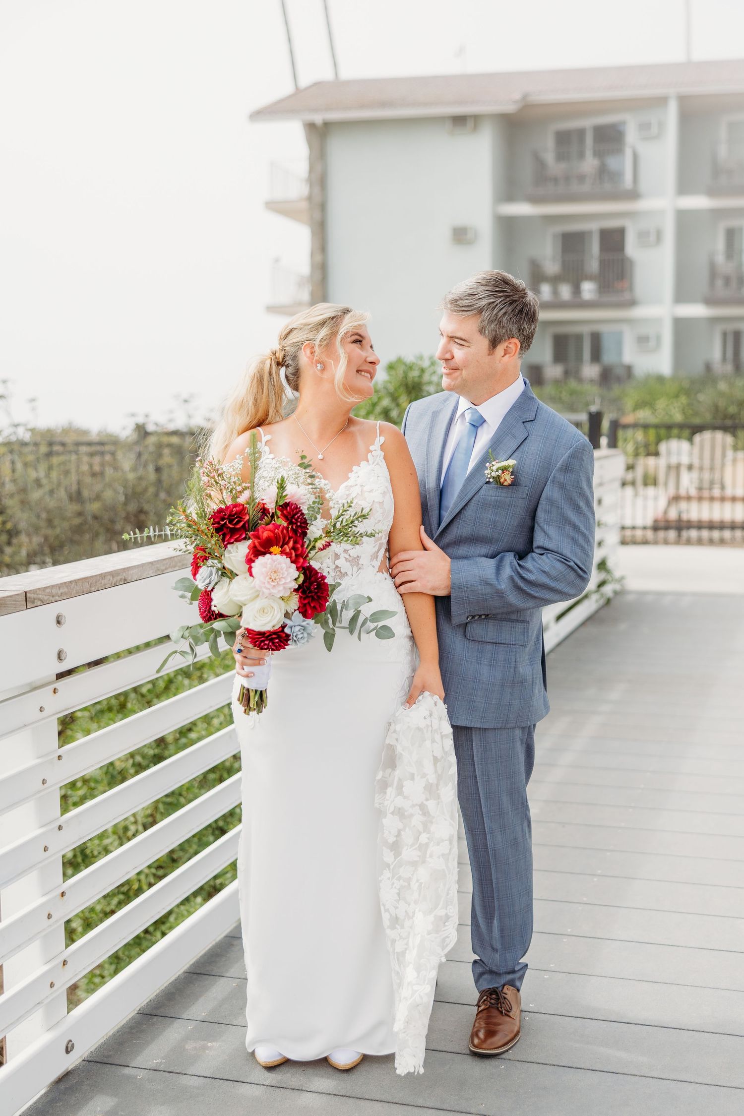 Newlyweds share a moment on beachfront deck with red and white floral bouquet.