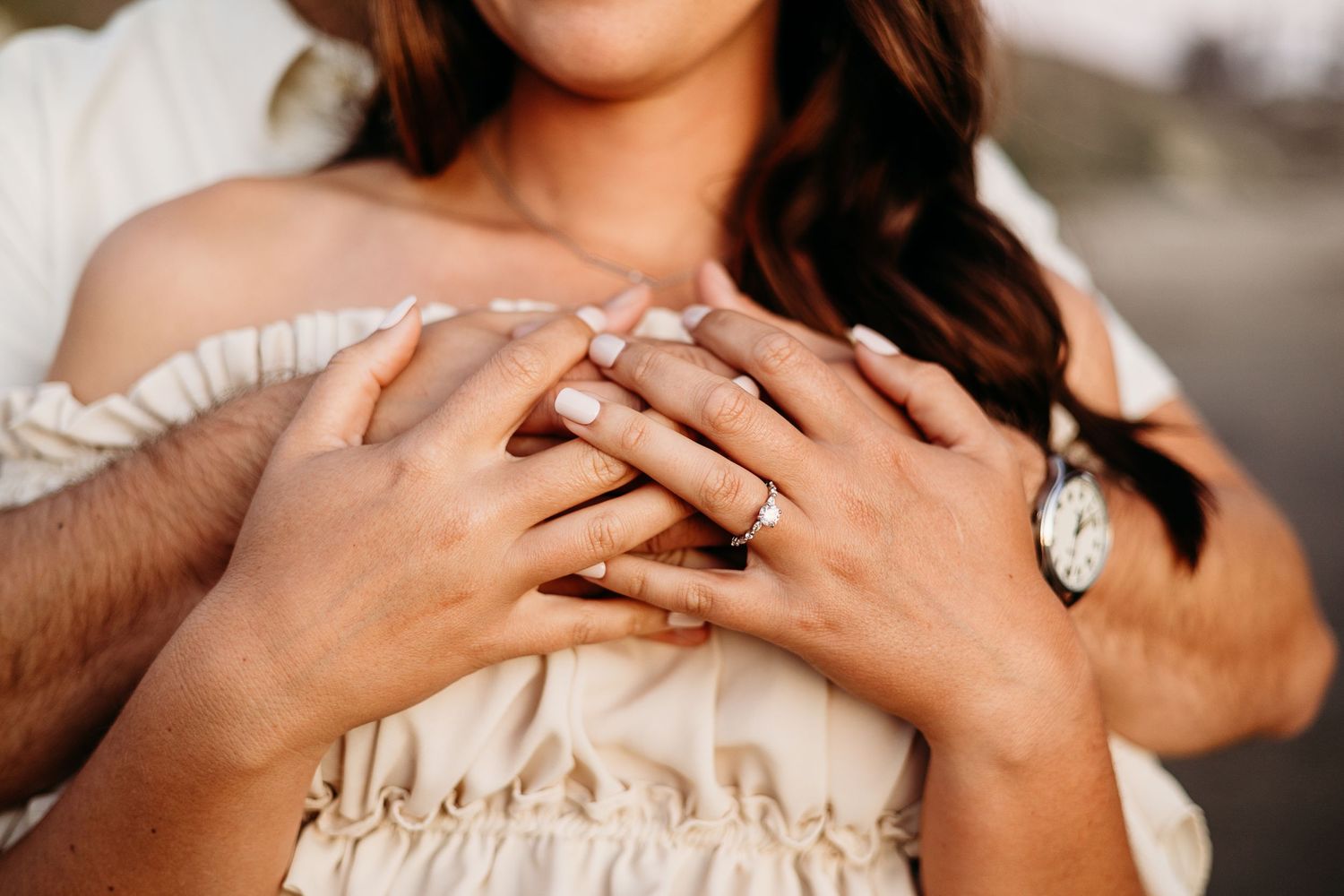 Close-up of hands displaying an engagement ring against a cream-colored dress.