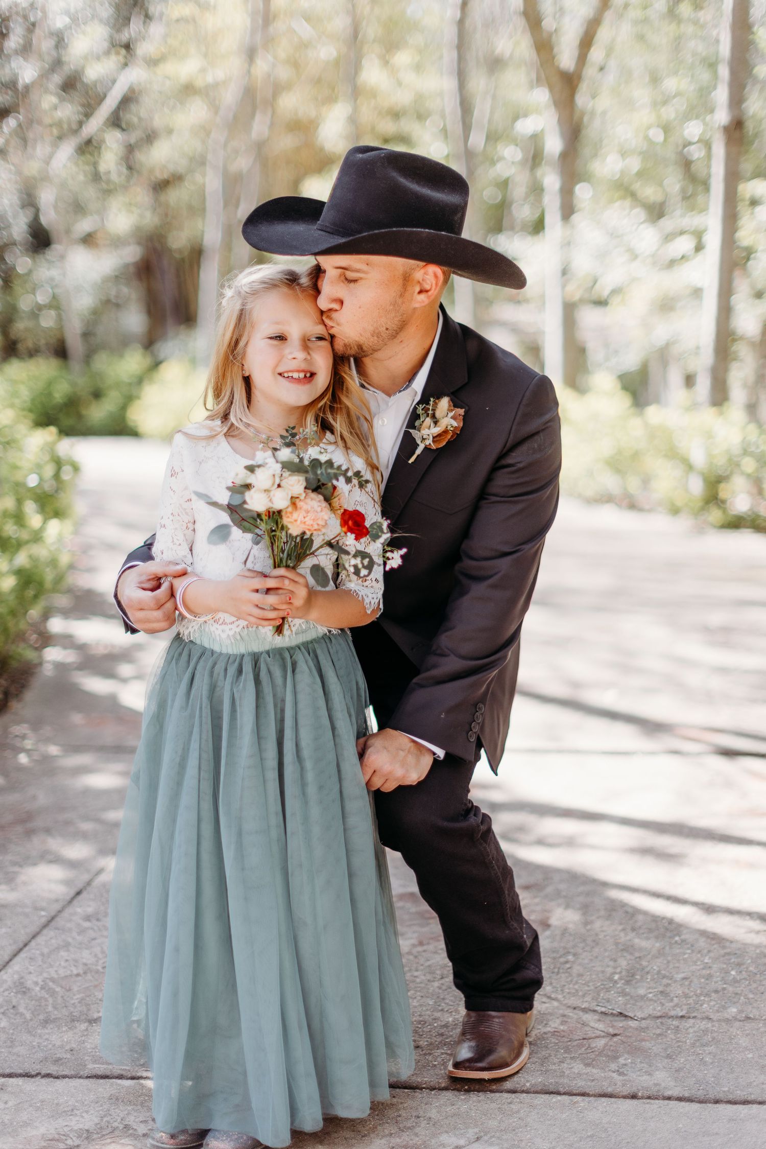 A person in a cowboy hat kisses on the cheek while holding a bouquet of flowers in an outdoor setting.