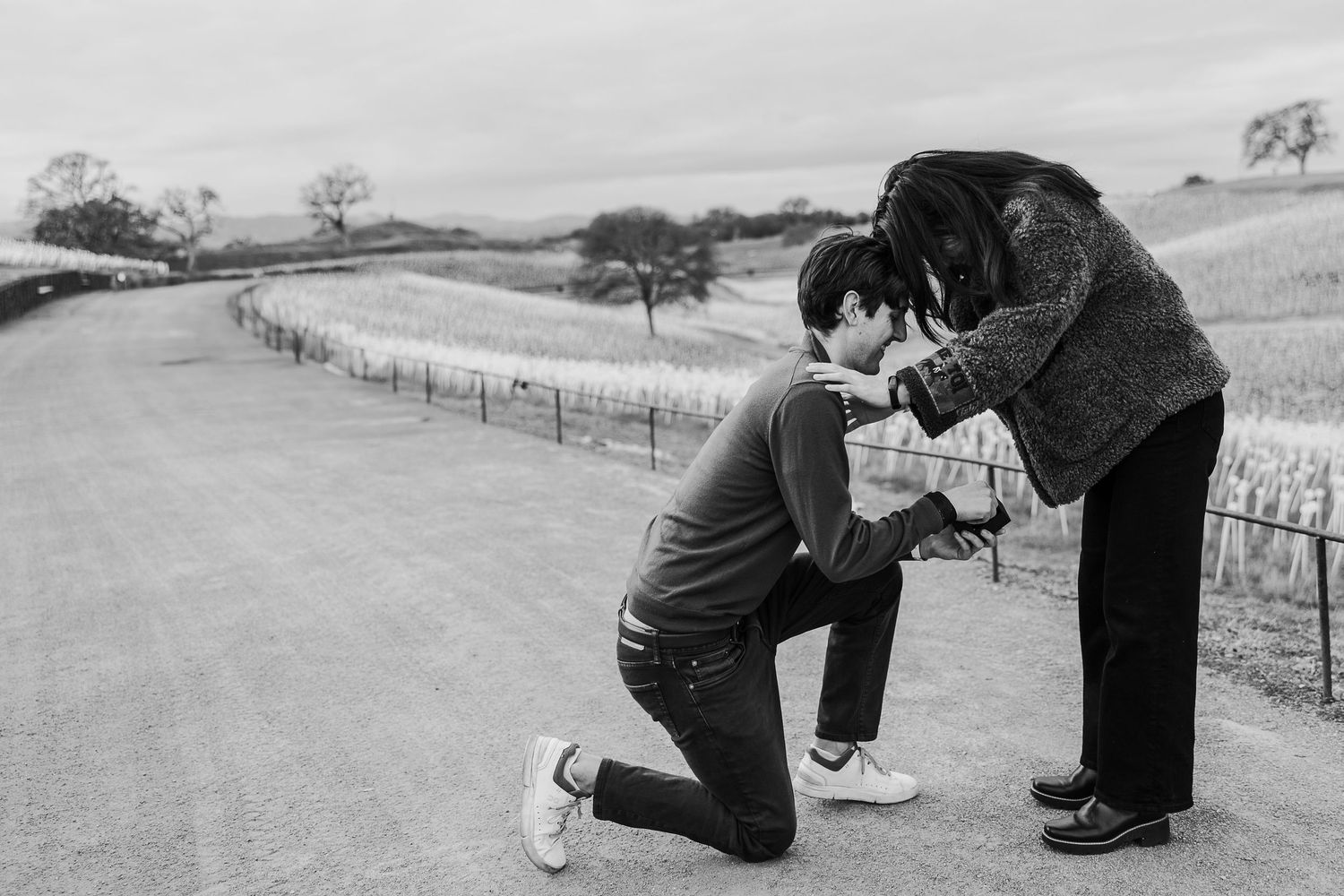 A tender black and white moment captured on a winding country road with a scenic rural landscape in the background.