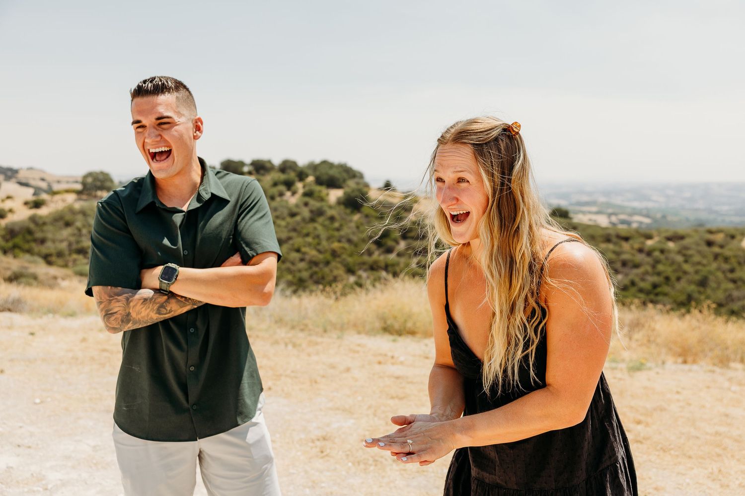 Couple sharing joyful moments while sitting in wooden chairs on a hilltop overlooking a scenic landscape.