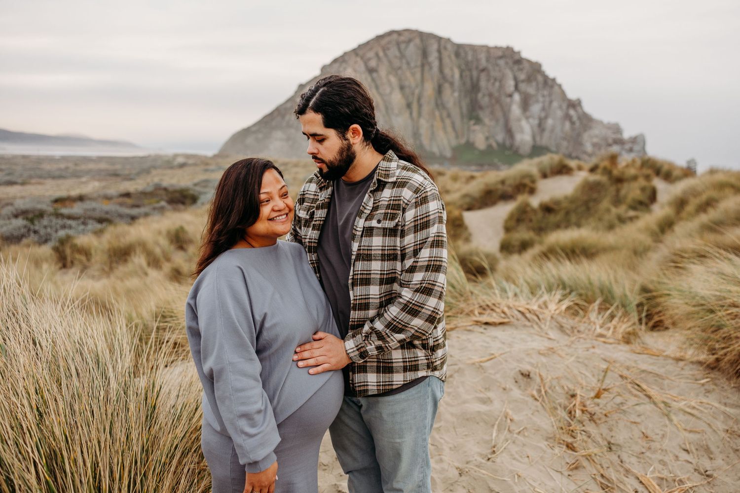 Couple embracing on sandy dunes with Morro Rock visible in background at sunset.