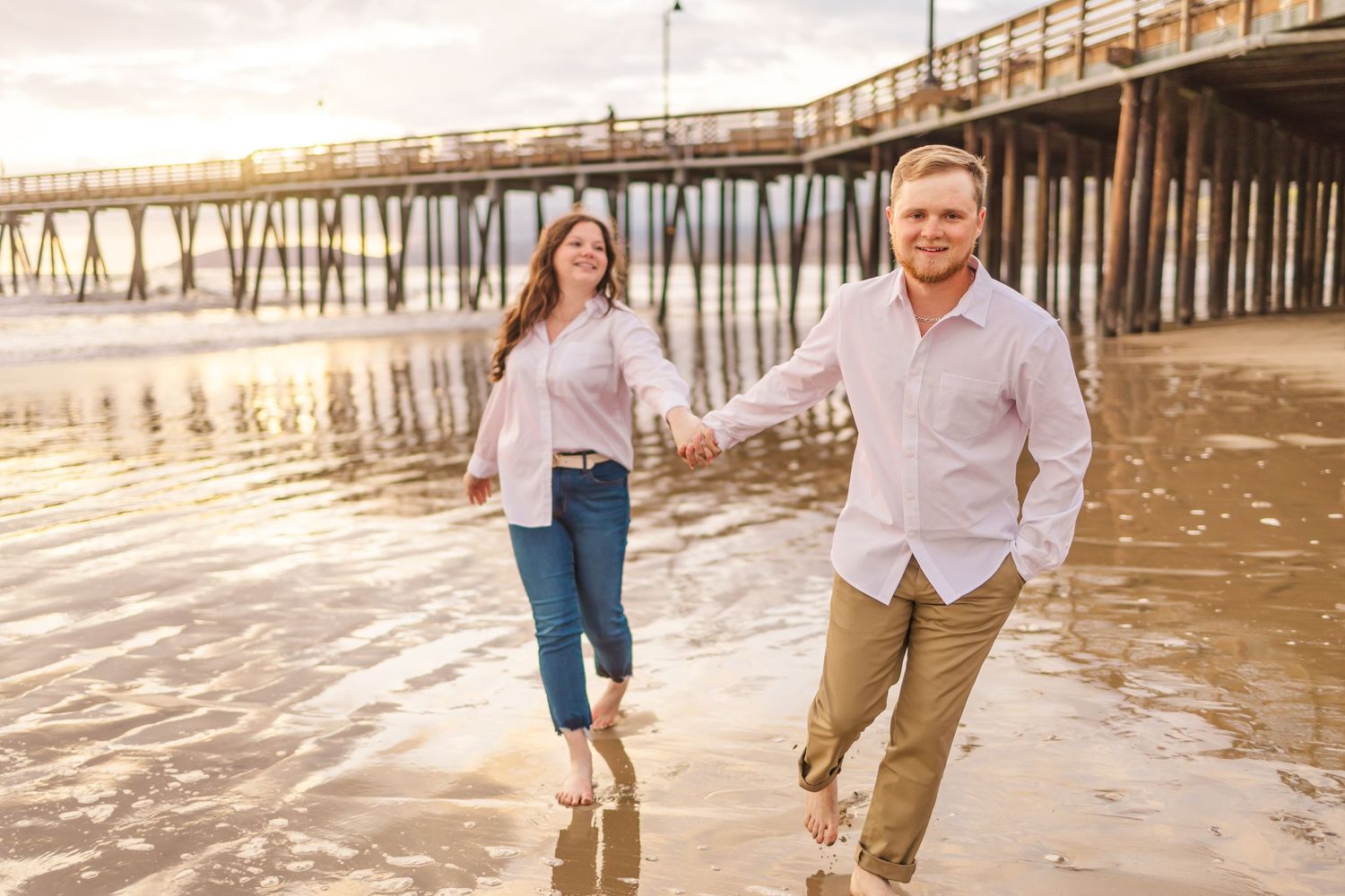 A couple runs joyfully along the beach at sunset with a wooden pier in the background.