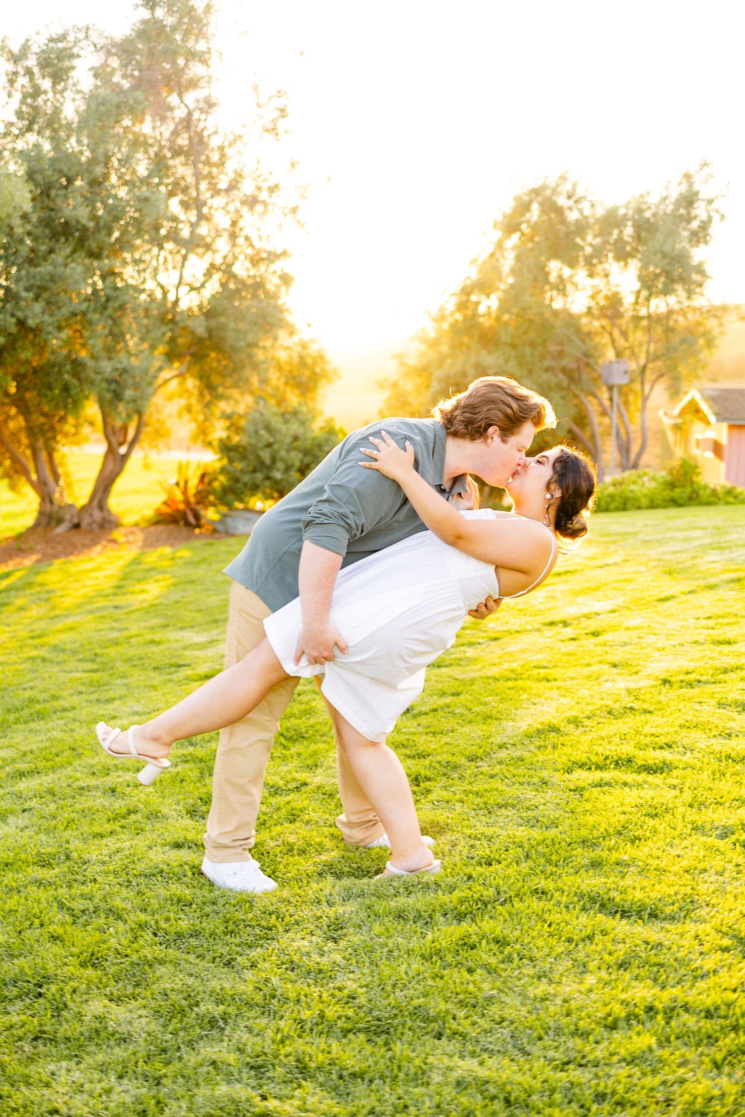 Playful couple dips during romantic photoshoot on sunny grass lawn.