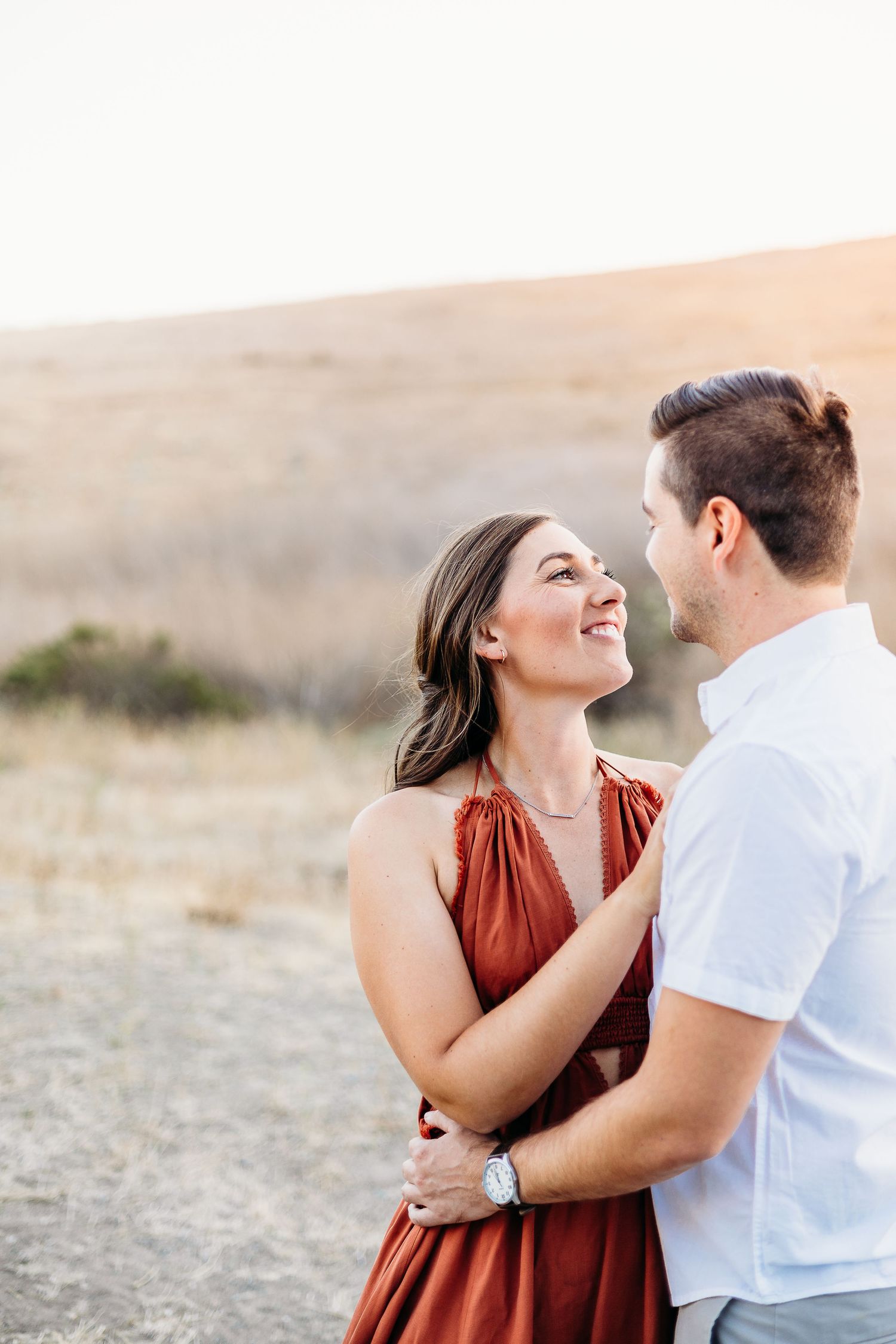 Couple in white shirt and rust dress share a loving moment in a natural outdoor setting.