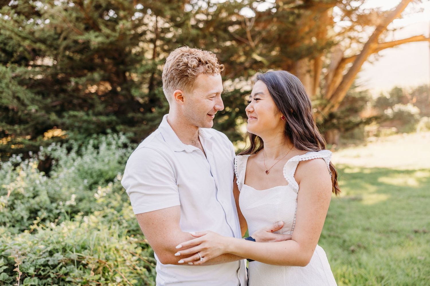 A couple laughs together in matching white outfits against a backdrop of summer foliage.