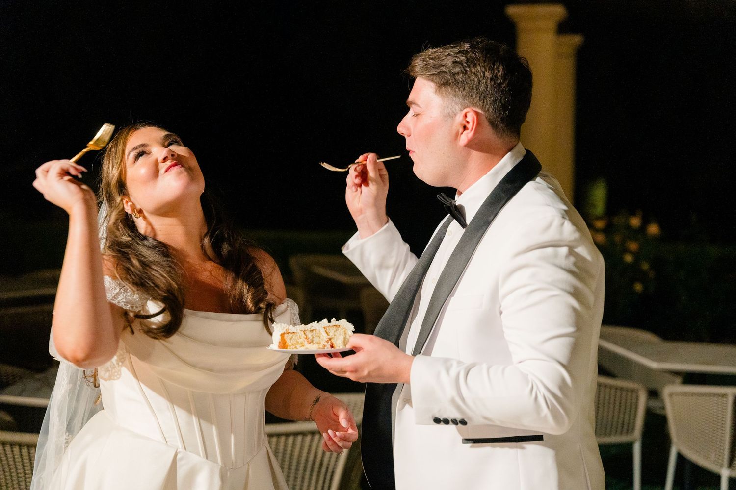 Bride and groom feeding each other cake under night sky at outdoor wedding reception.