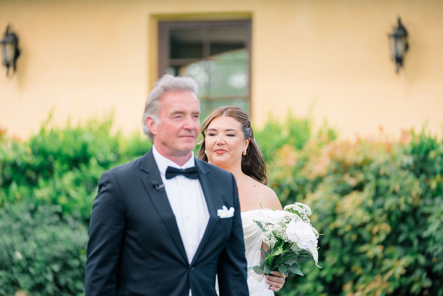 Bride and her father walk together outdoors before wedding ceremony, wearing white gown and black tuxedo.