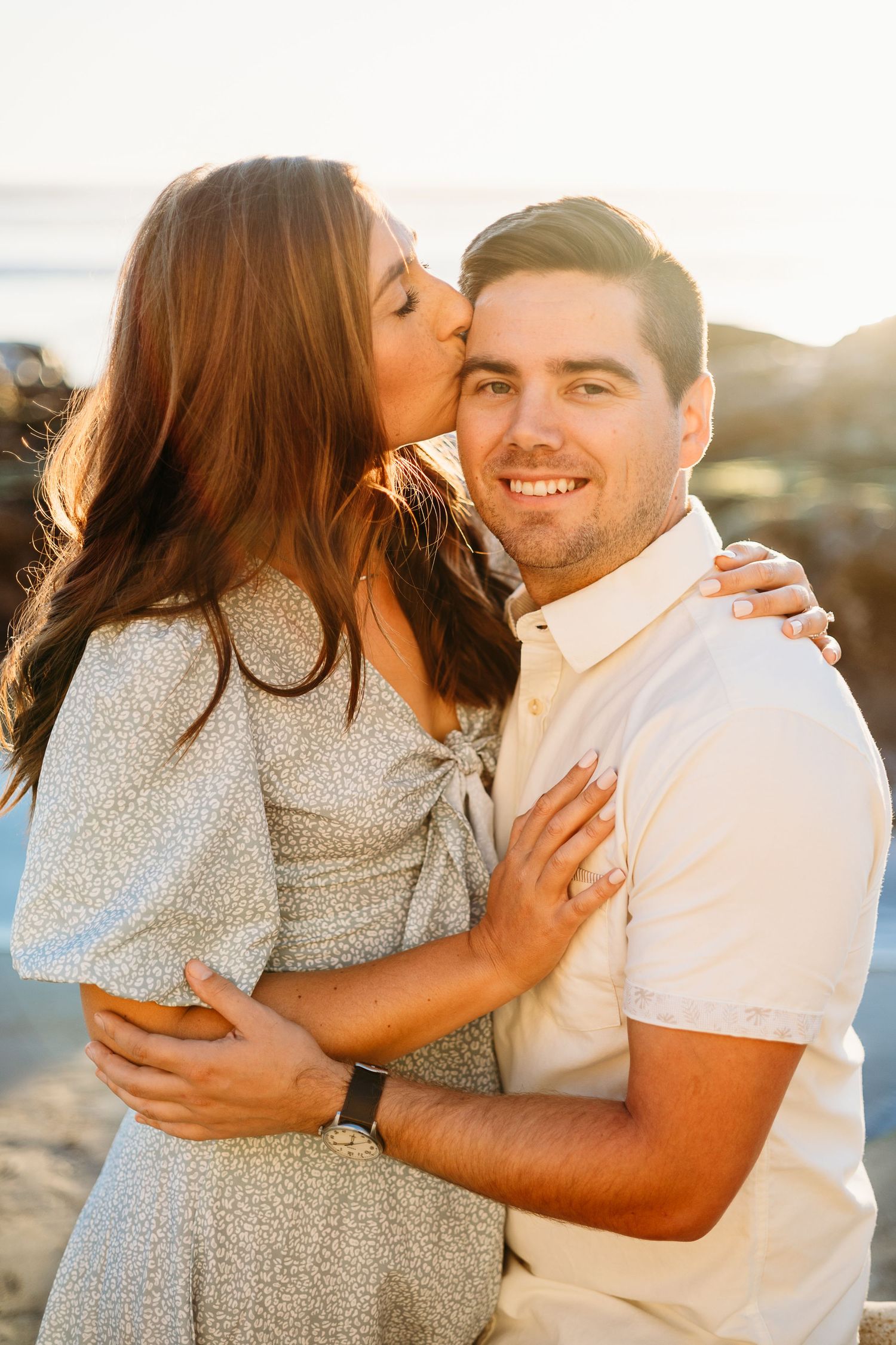 A romantic couple embraces during golden hour at the beach as she kisses his cheek.