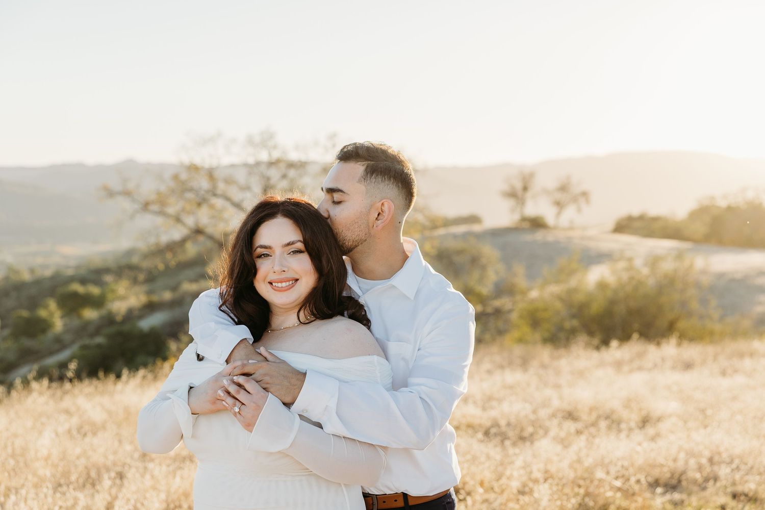 A couple embraces in a romantic moment on a sunlit hilltop with golden grass and rolling hills in the background.