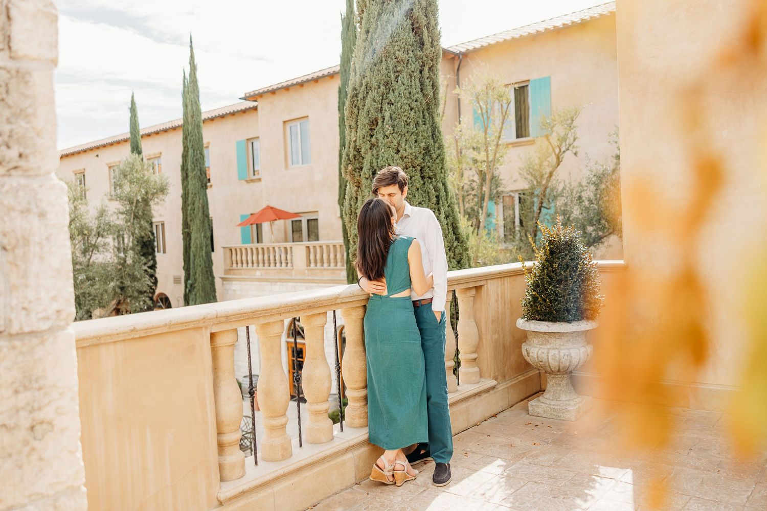 A couple standing on a sunny balcony with Mediterranean architecture and cypress trees in the background.