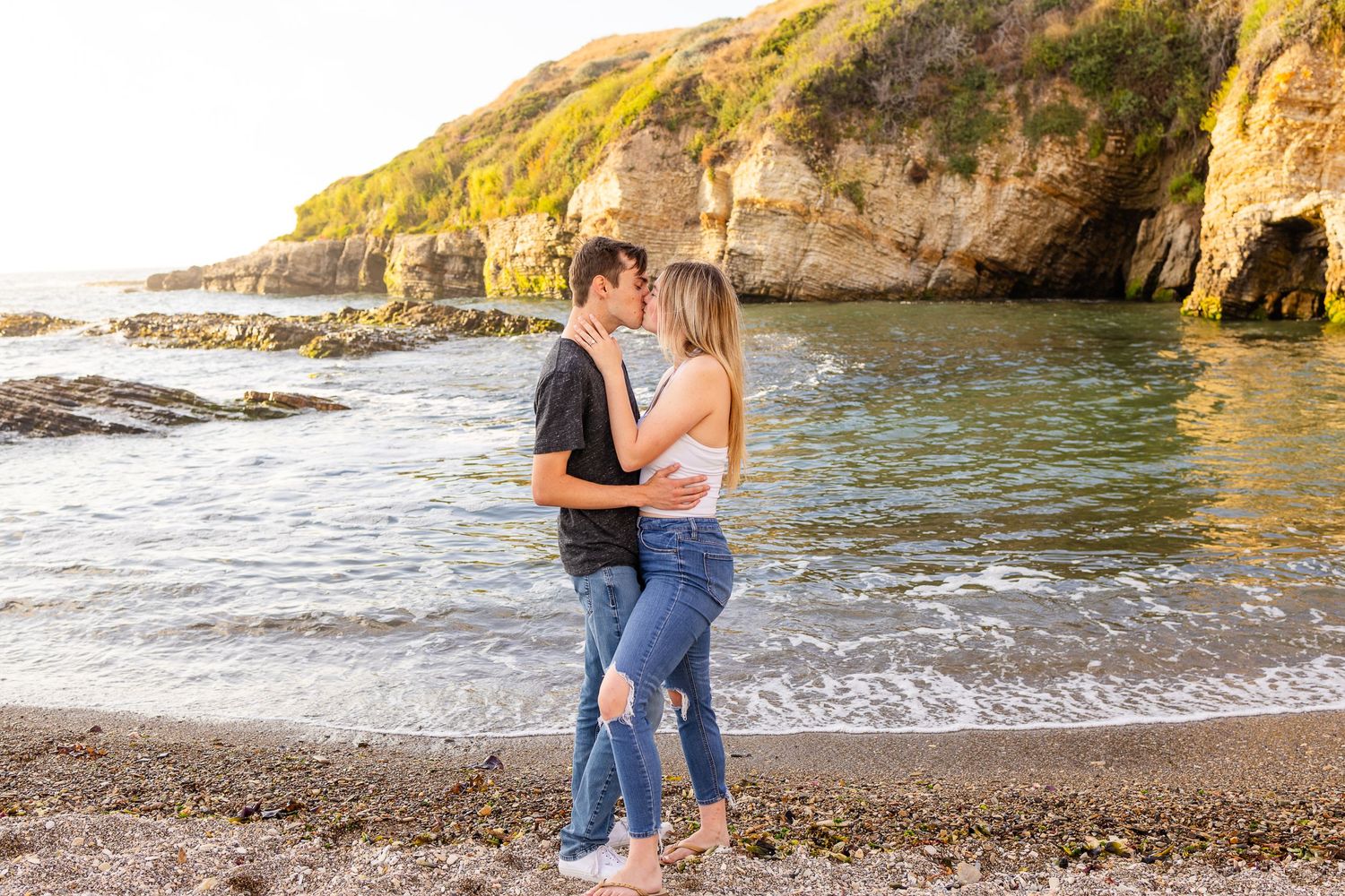 A couple embraces on a scenic beach with cliffs and ocean waves in the background.