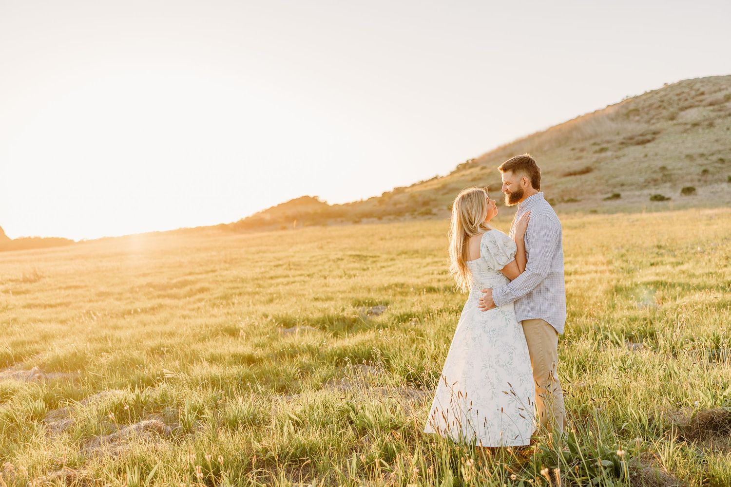 A couple embraces in a golden field at sunset with mountains in the background during a romantic moment.
